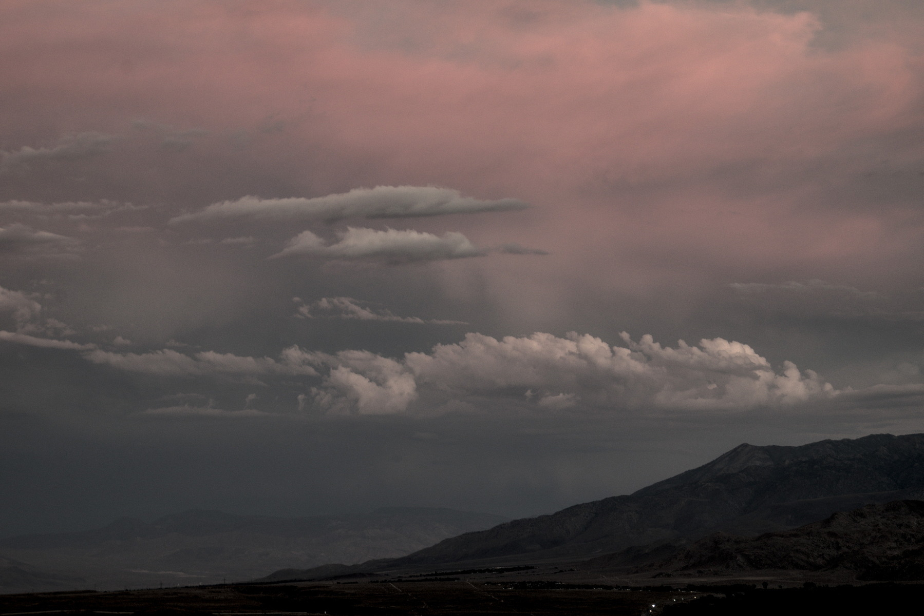 A narrow line of foft grey clouds float over the trailing edge of a mountain range, with pink skies behind.
