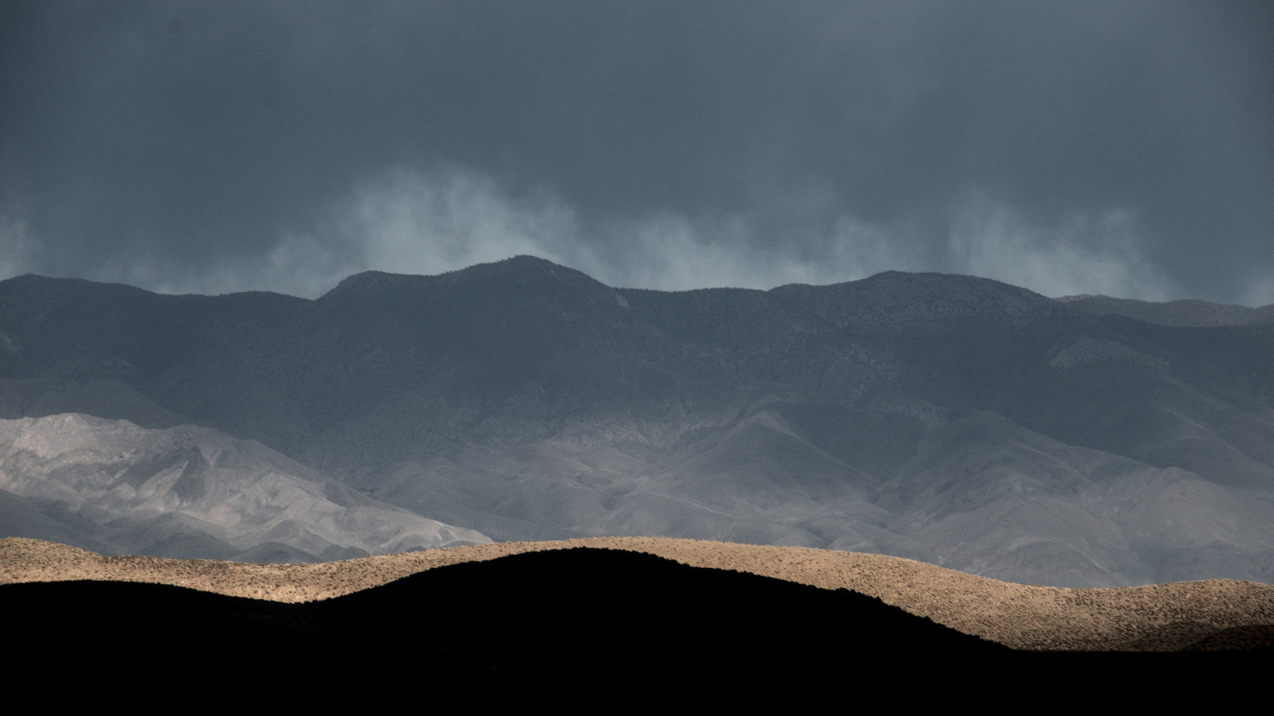 Hills receding to the White Mountains, black shadow in the foreground, blue, grey rain in the background, with a thin line of sage brush lit up golden in the middle.