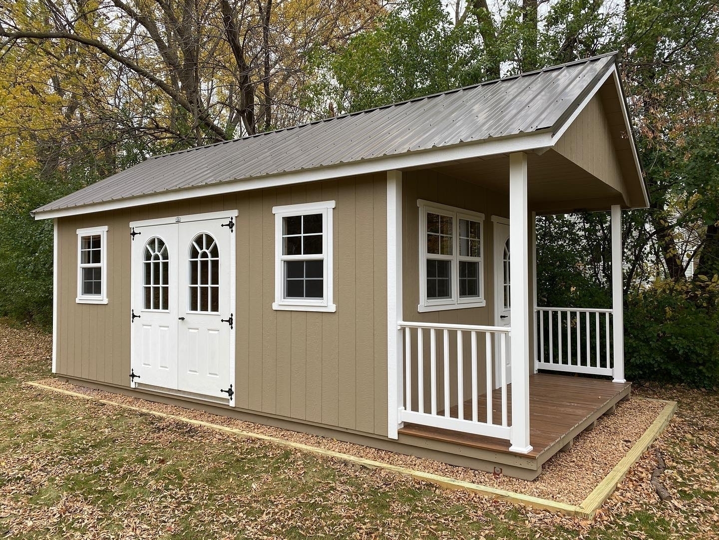 A small, beige shed with white-trimmed windows and arched double doors stands on a wooden base. Surrounded by autumn trees and fallen leaves, it features a small porch.