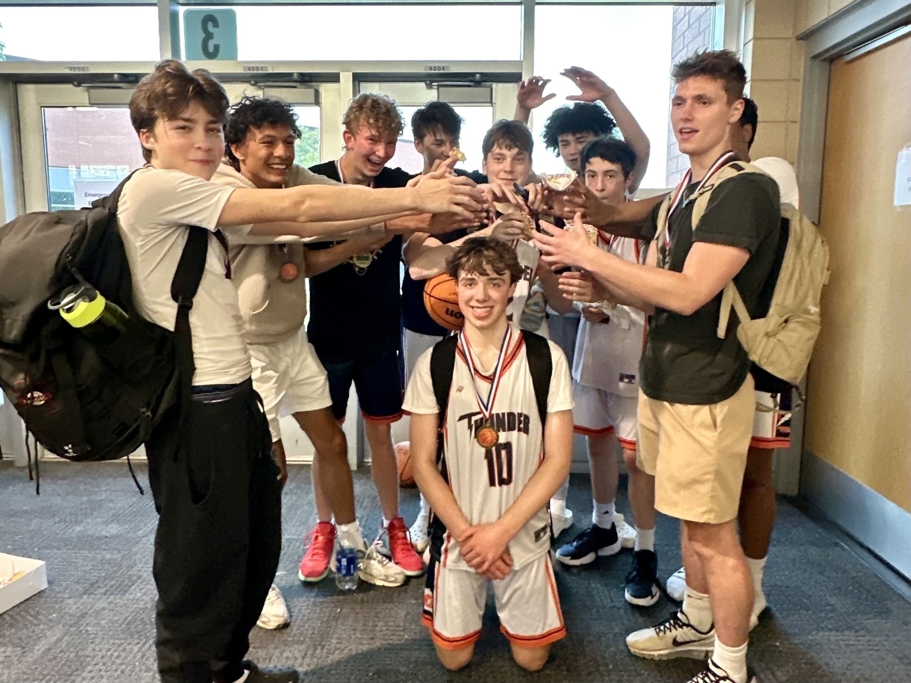 Auto-generated description: A group of young basketball players celebrates with a trophy in a hallway, some wearing medals and uniforms
