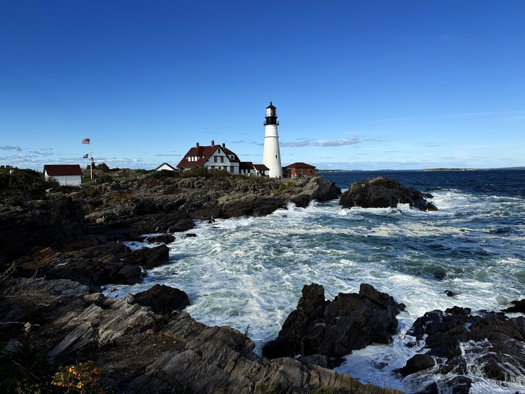 A lighthouse stands on rocky cliffs beside crashing ocean waves, with houses and an American flag in the background under a clear blue sky.