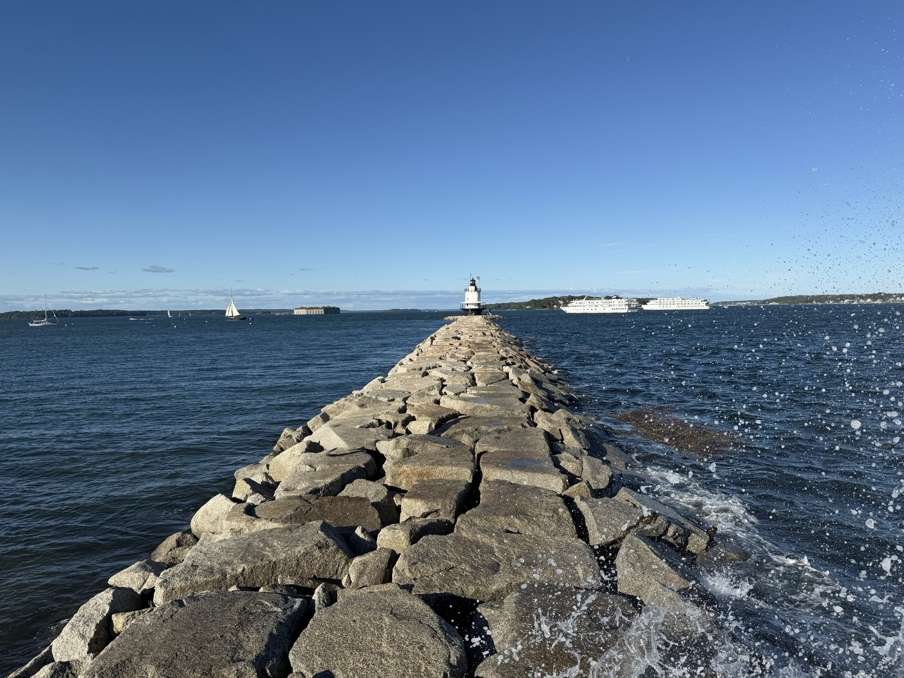 A stone pathway extends into the ocean, leading to a small lighthouse. Sailboats and two large vessels float on the water under a clear blue sky.