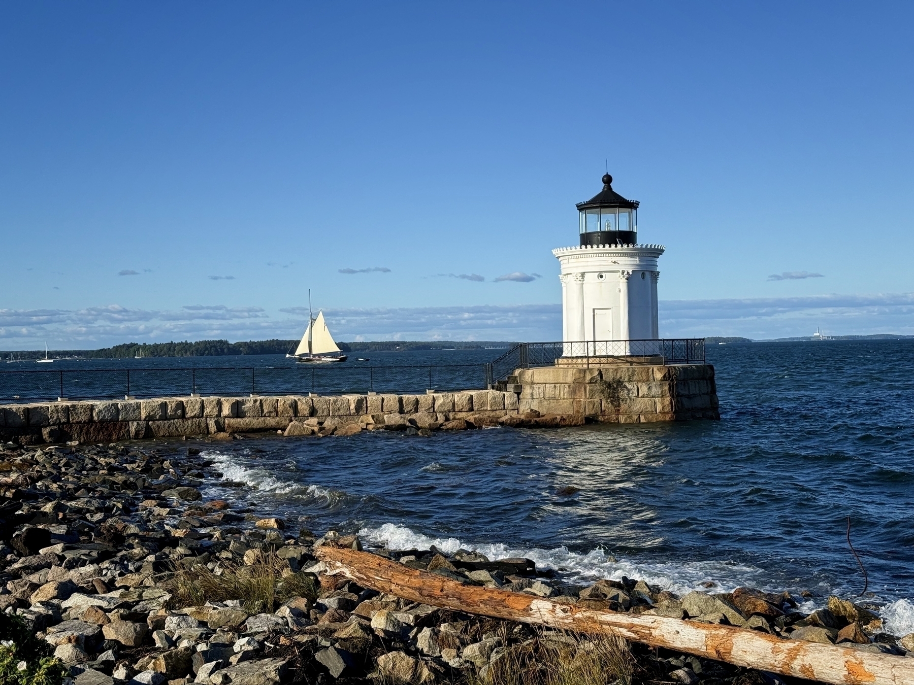 A white lighthouse stands on a rocky shore beside a calm, blue sea, with a sailboat gliding in the background under a clear sky.