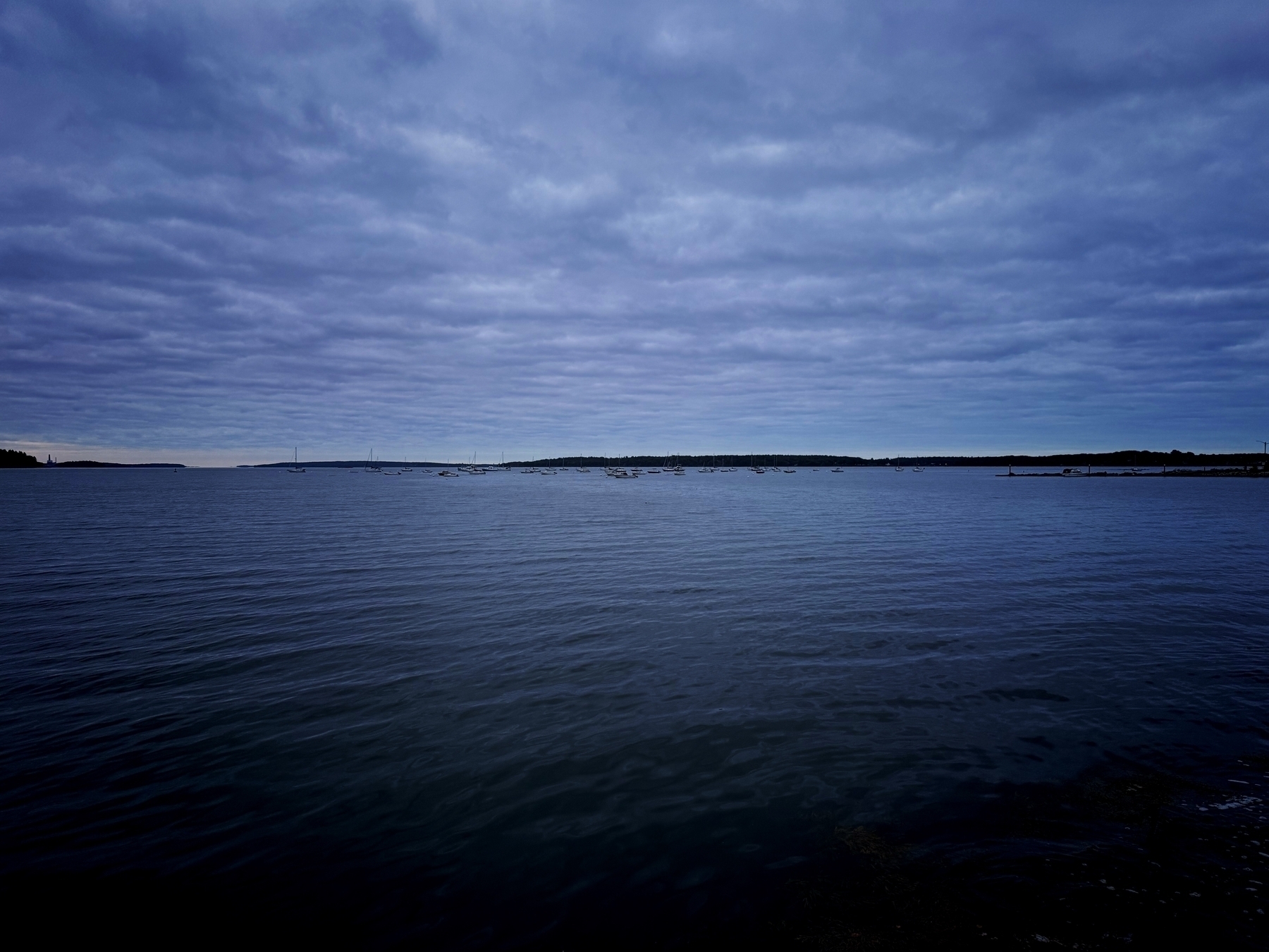 A calm sea under a cloudy sky with a distant shoreline visible, scattered with small boats on the horizon.