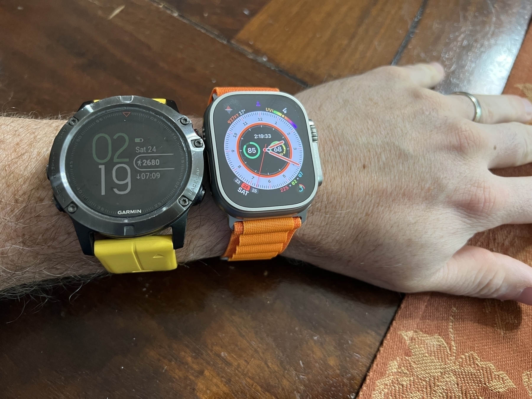 Two smartwatches on a wrist display different time and fitness information, resting on a wooden surface next to a patterned cloth.