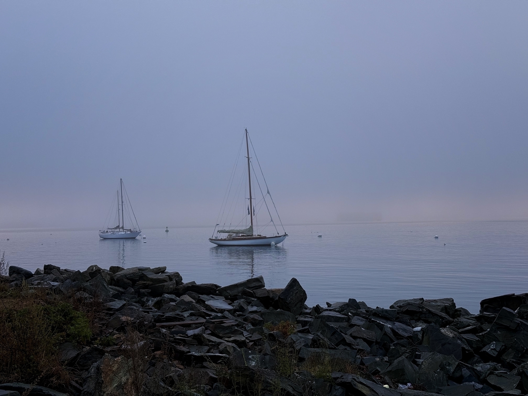 Two sailboats float on calm water with scattered buoys, surrounded by a rocky shoreline, under a dense, foggy sky.