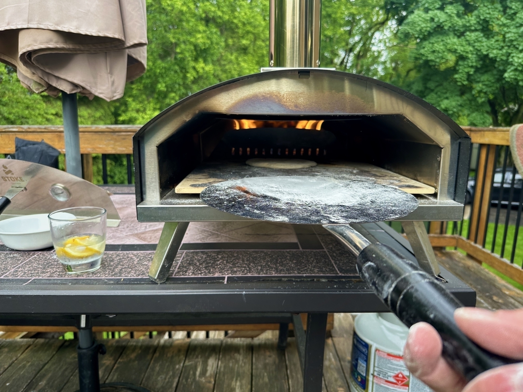 A pizza oven flames inside on a wooden table outdoors with a spatula lifting dough nearby. Items like a glass with lemon slices and an umbrella are on the table.