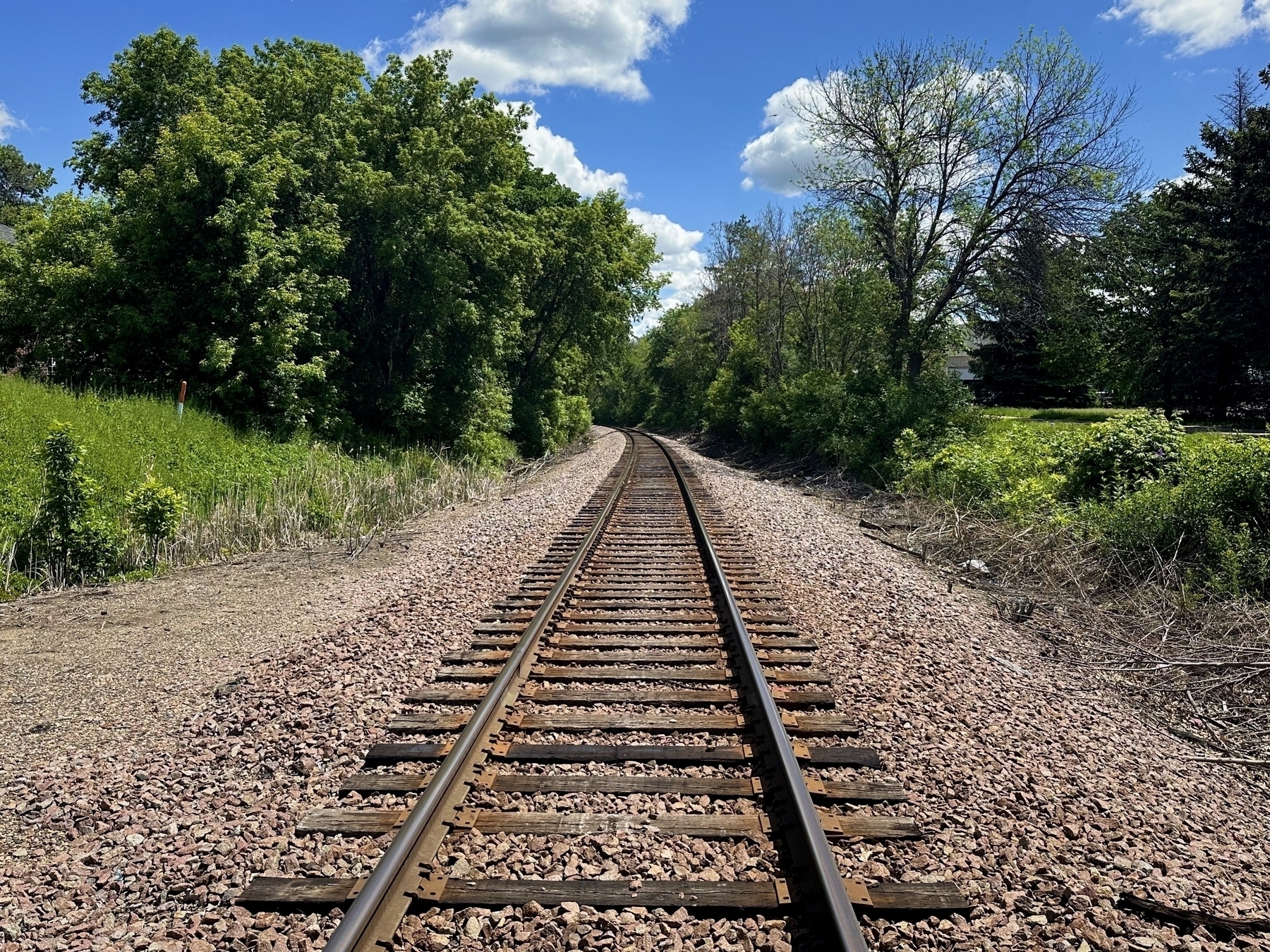 Railroad tracks stretch into the distance surrounded by trees and greenery beneath a blue sky with scattered clouds.