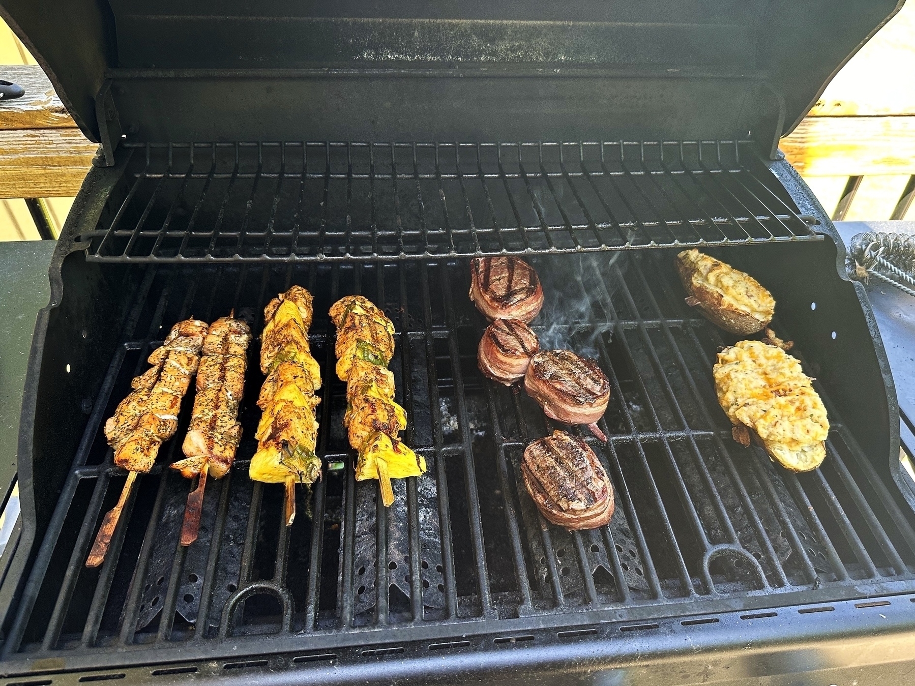Various grilled foods are cooking on an open outdoor grill including skewered meat vegetable kebabs steak medallions wrapped in bacon and twice-baked potatoes