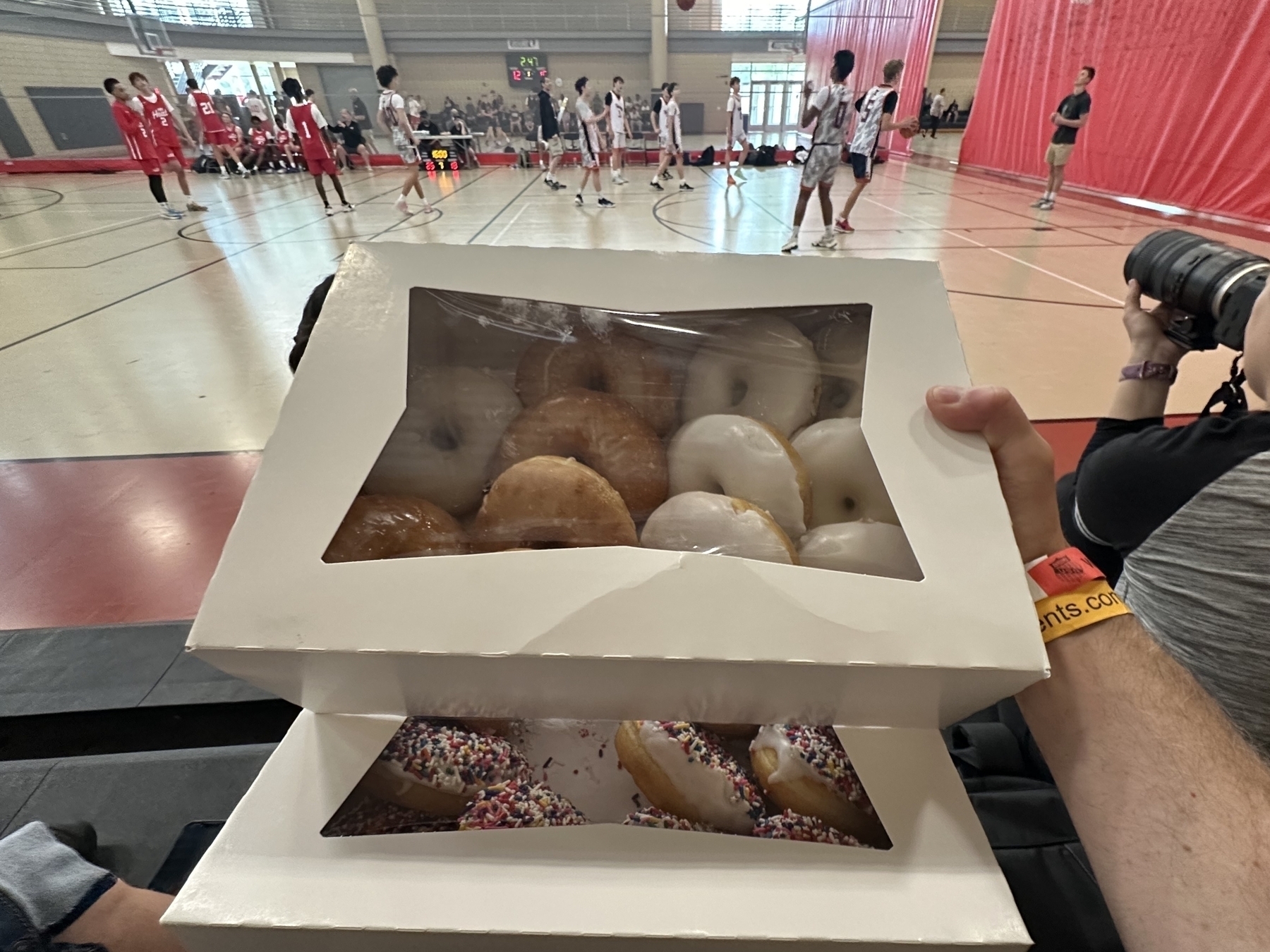 Two boxes of assorted donuts are held by a person in a gym where a basketball game is occurring between teams in red and white uniforms.