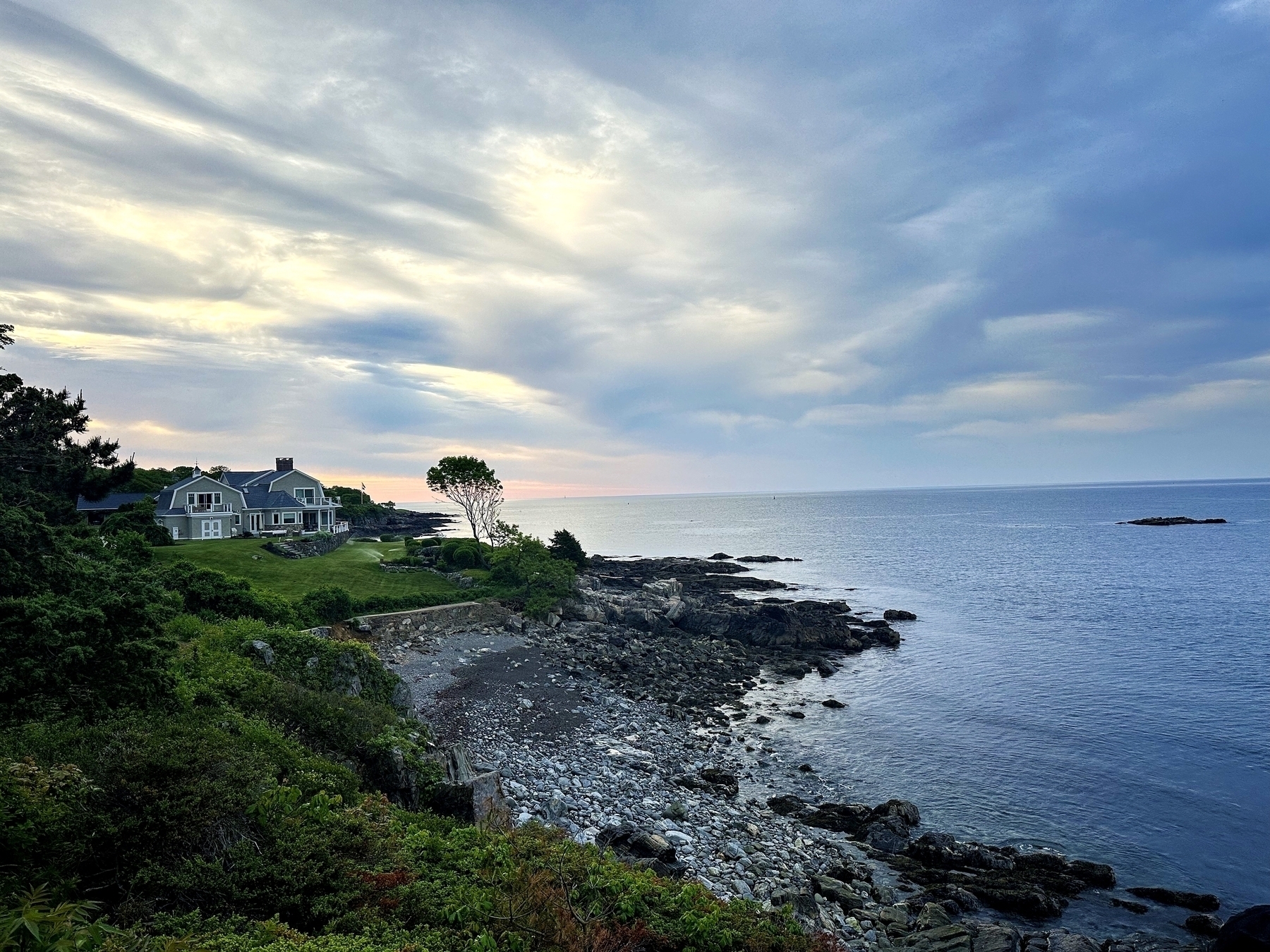 A house overlooks a rocky shoreline beside a calm ocean under a partly cloudy sky with blue and white hues.