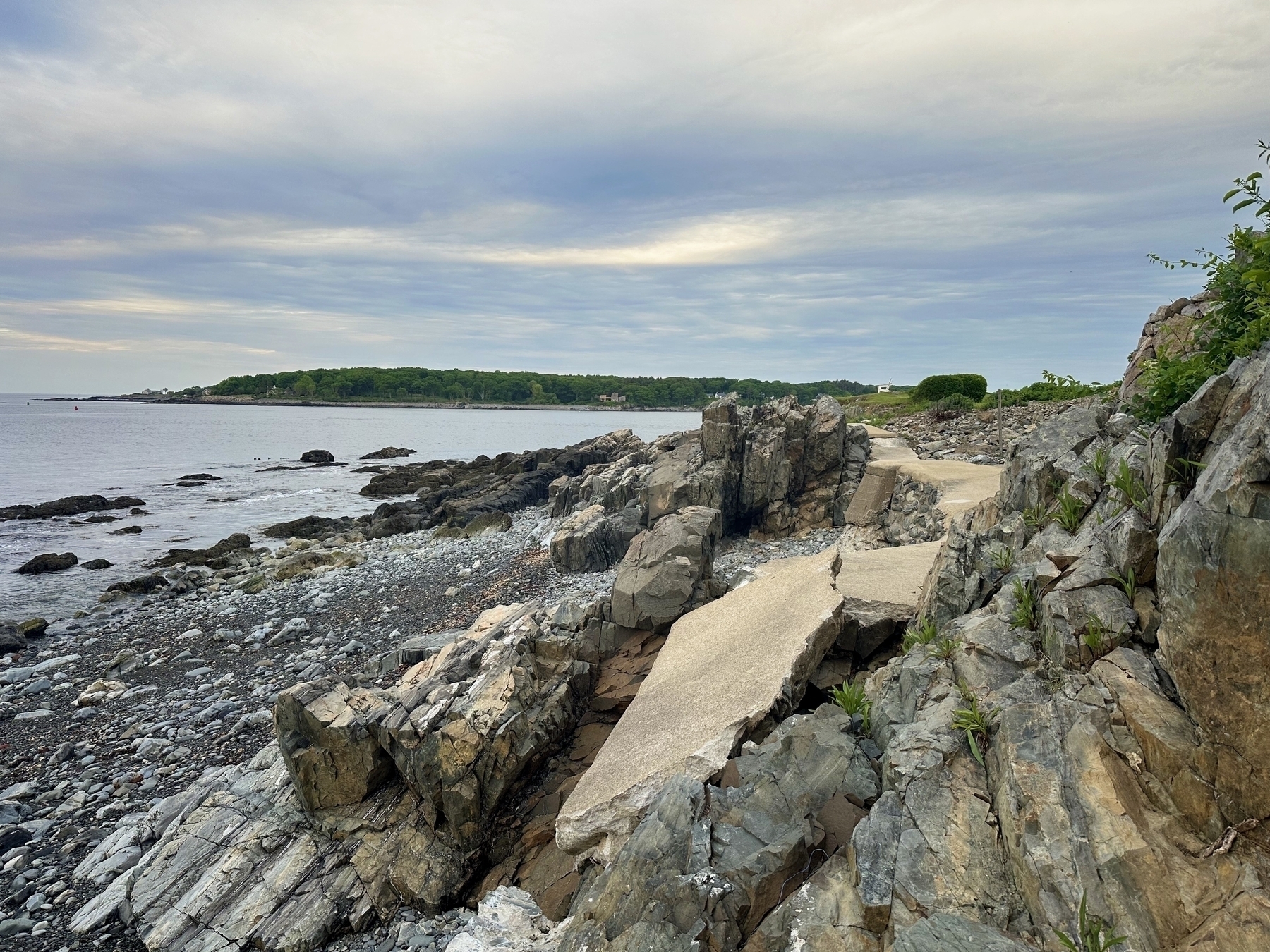 Rocky coastline adjacent to calm sea under a cloudy sky with a concrete pathway winding through large rocks and distant greenery displayed on the horizon.