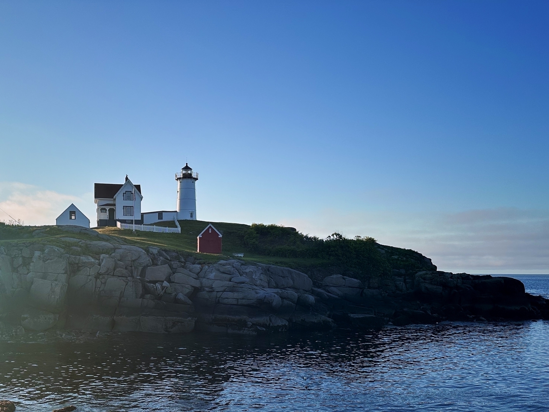 A lighthouse with an attached house and outbuildings stands on a rocky cliff overlooking a calm ocean under a clear blue sky.
