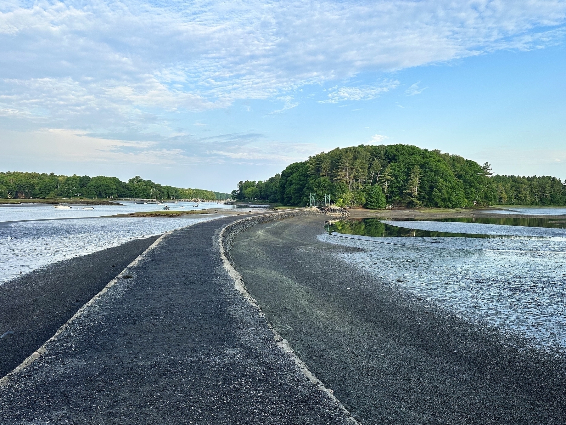 A narrow paved path stretches towards a tree-covered island surrounded by low tide waters under a partly cloudy sky