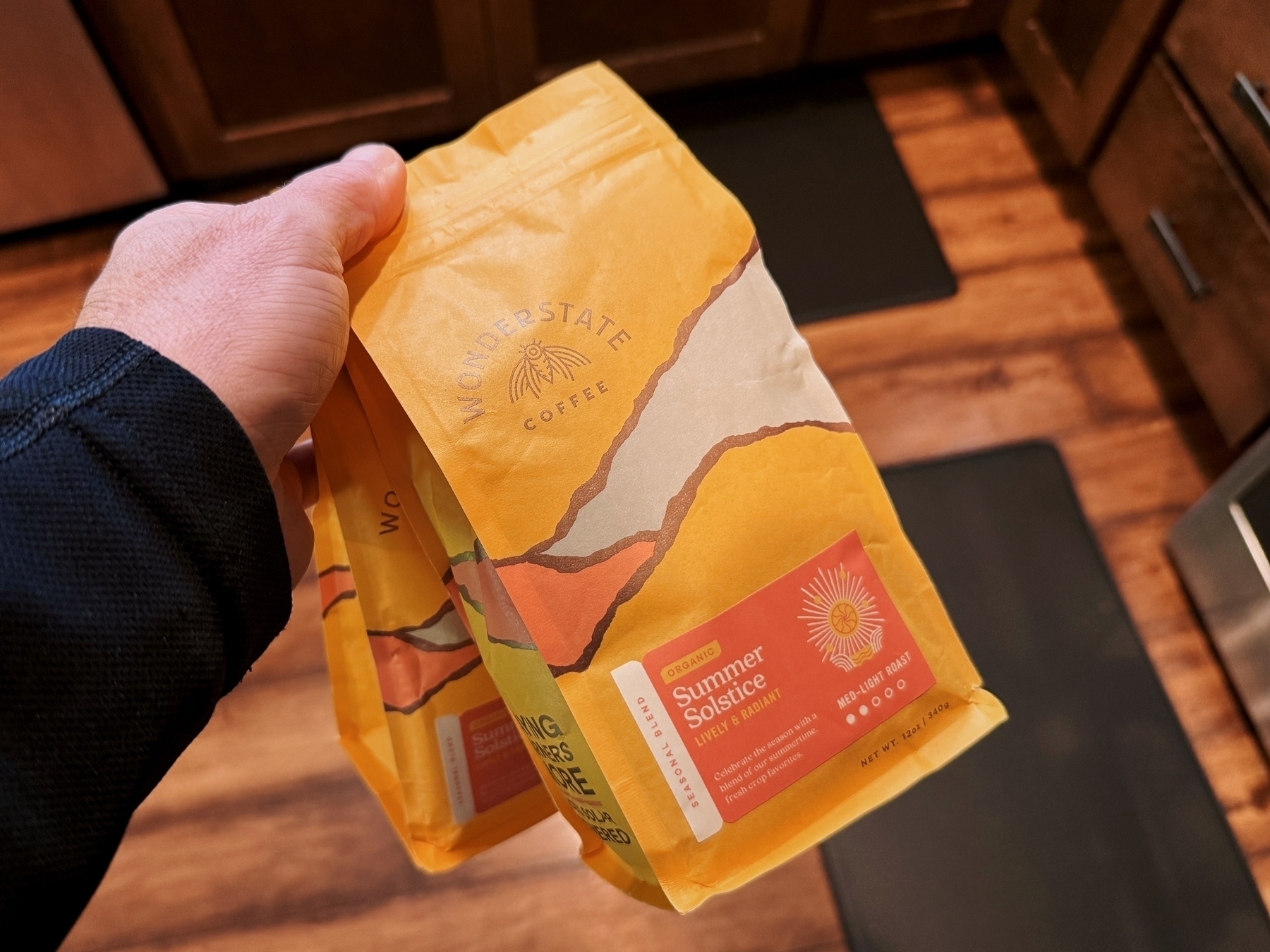 A hand holds a yellow coffee bag labeled “Wonderstate Coffee - Summer Solstice, Organic, Seasonal Blend, Med-Light Roast.” The setting is a wooden-floored kitchen with visible cabinets.