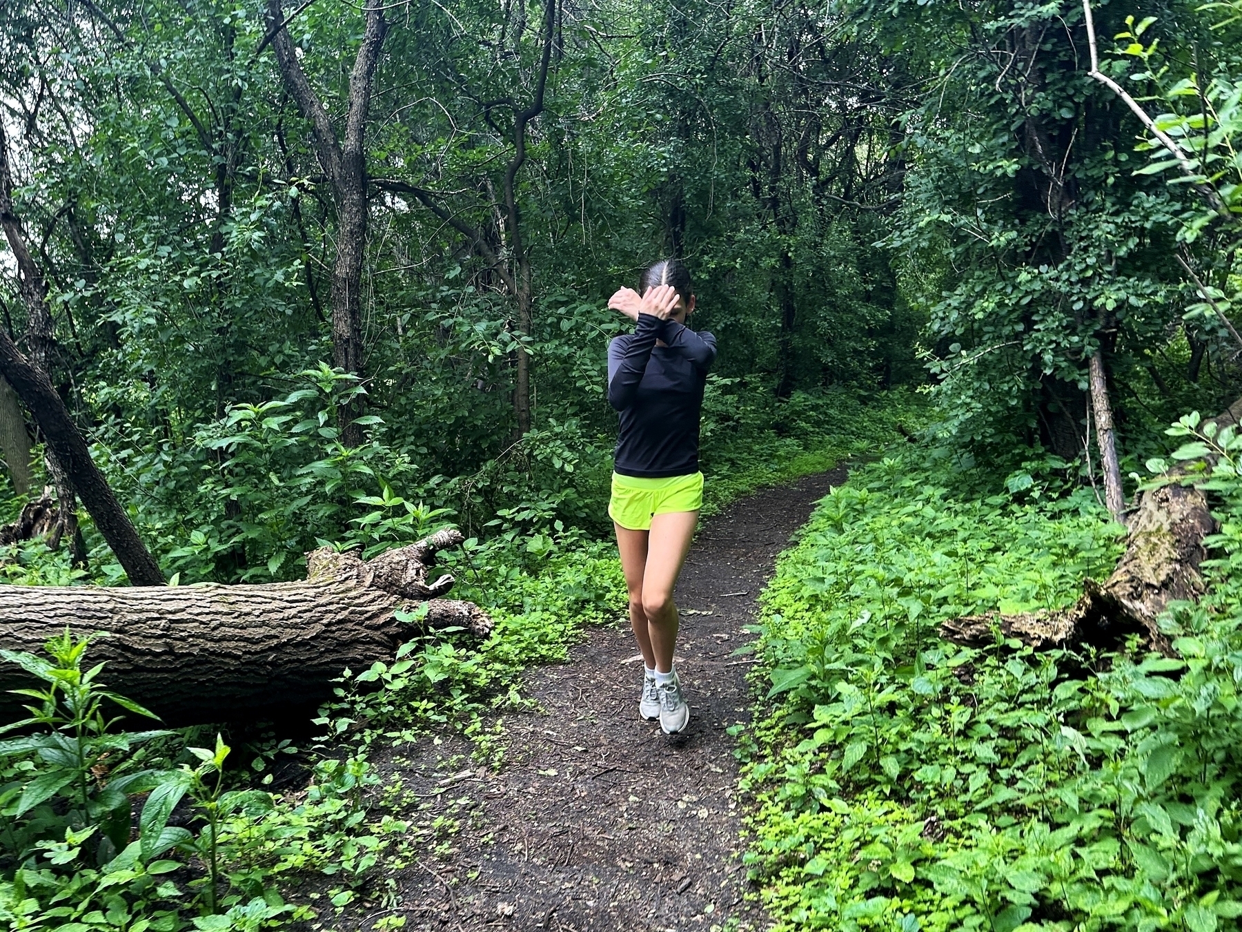 Person running through a dense forest with leafy, green plants and trees; the person is wearing a black top and neon yellow shorts and is covering their face with their hands.