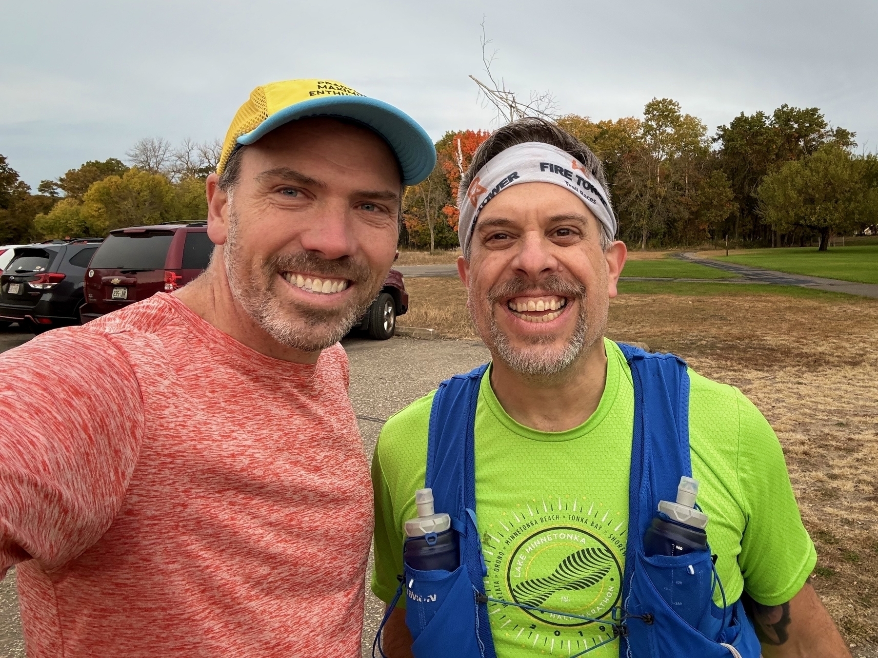 Two men are smiling for a selfie outdoors, wearing athletic gear and standing in a park with trees and cars in the background.