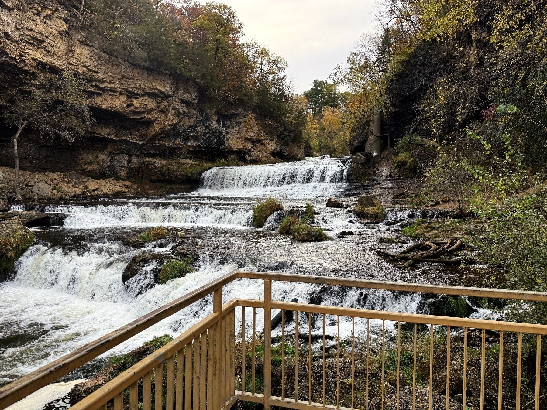 A multi-tiered waterfall cascades down rocky ledges surrounded by autumn foliage and is viewed from a wooden observation deck.