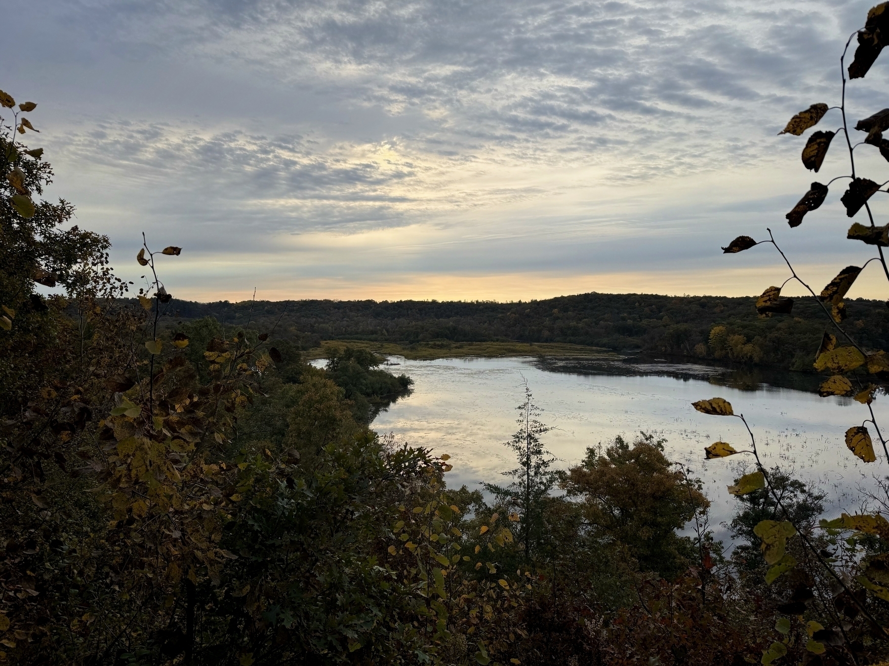 River winds through a forested landscape, reflecting the cloudy sky. Trees with autumn leaves frame the foreground, creating a serene, natural setting at dusk.