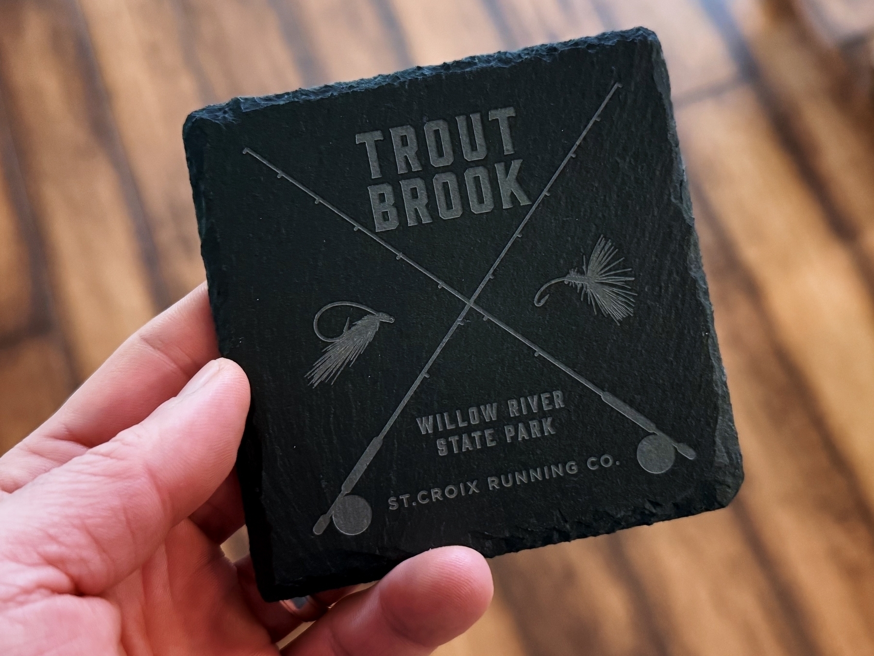 A hand holds a square, dark slate coaster featuring crossed fishing rods, flies, and text: “TROUT BROOK” and “WILLOW RIVER STATE PARK” with “ST. CROIX RUNNING CO.” underneath. A wooden floor is visible in the background.