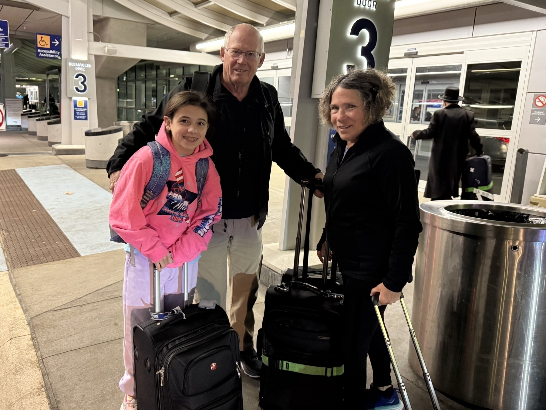 Three people stand together, smiling, each holding luggage by a doorway labeled “3” at an airport curbside.