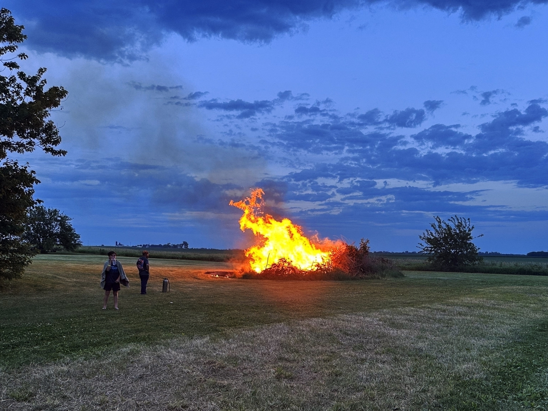 A blazing bonfire illuminates an open grassy field during twilight two people stand near the fire one watching and the other taking a photo with trees and a blue sky in the background