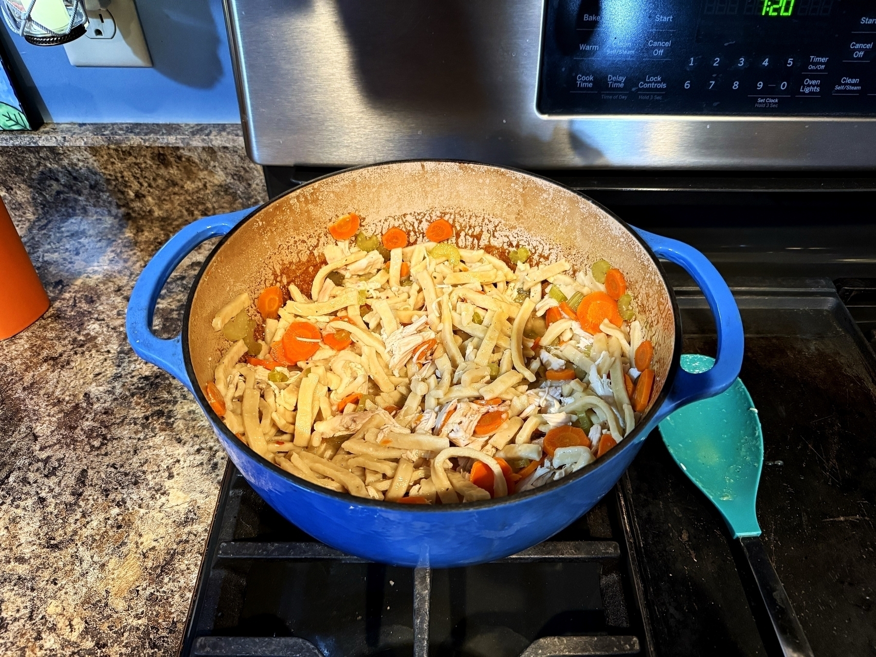 A blue pot on a stovetop contains cooked noodles, sliced carrots, and other vegetables. It sits on a kitchen counter beside a bright orange utensil holder and a digital clock.