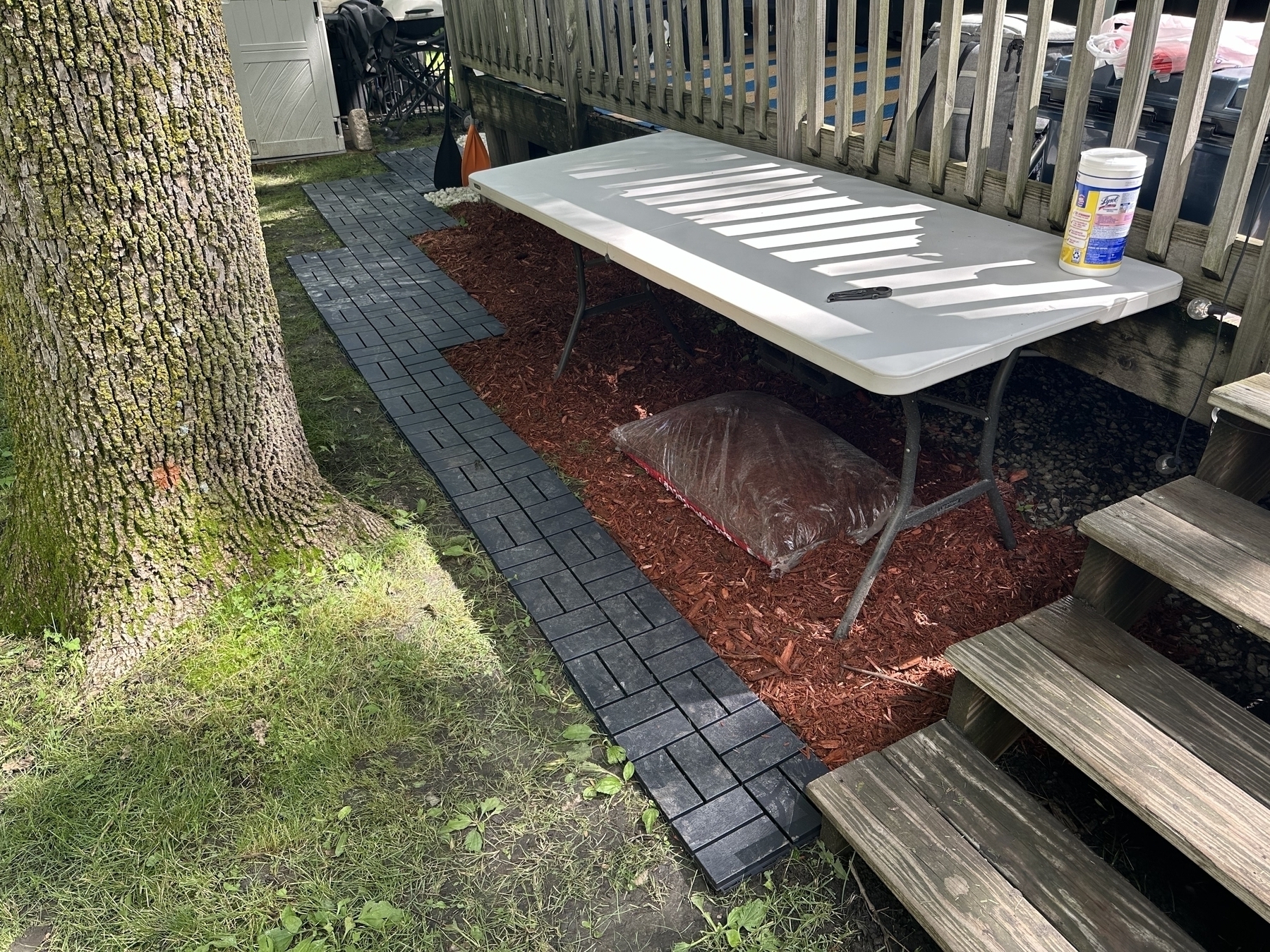 Plastic folding table with items on top including a canister placed alongside garden mulch bag beneath in an outdoor setting next to a patio deck and large tree.
