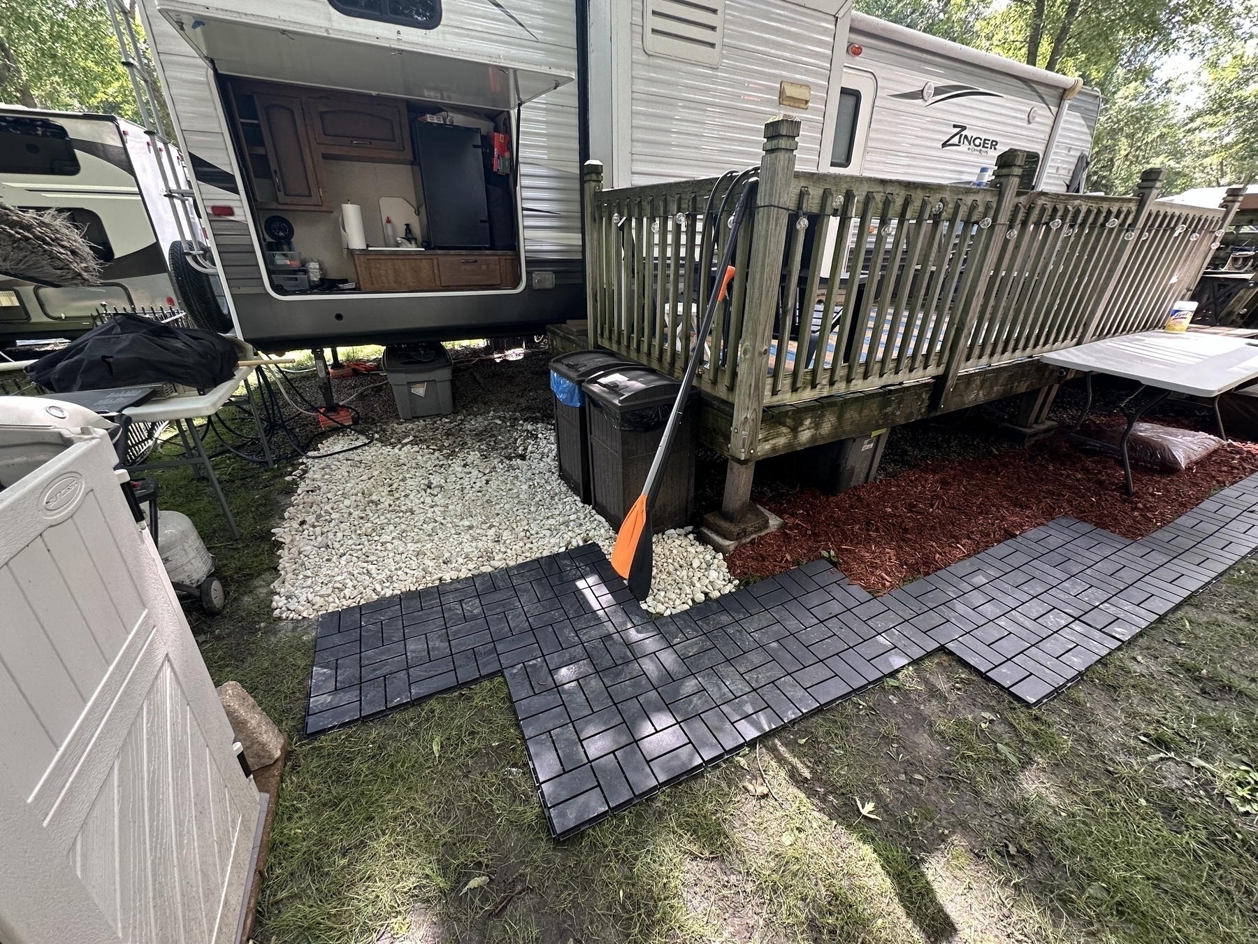 RV with an open side panel reveals a kitchen. A wooden deck fitted to the RV is surrounded by gravel and black tiles. Trees provide shade in the surrounding campground.