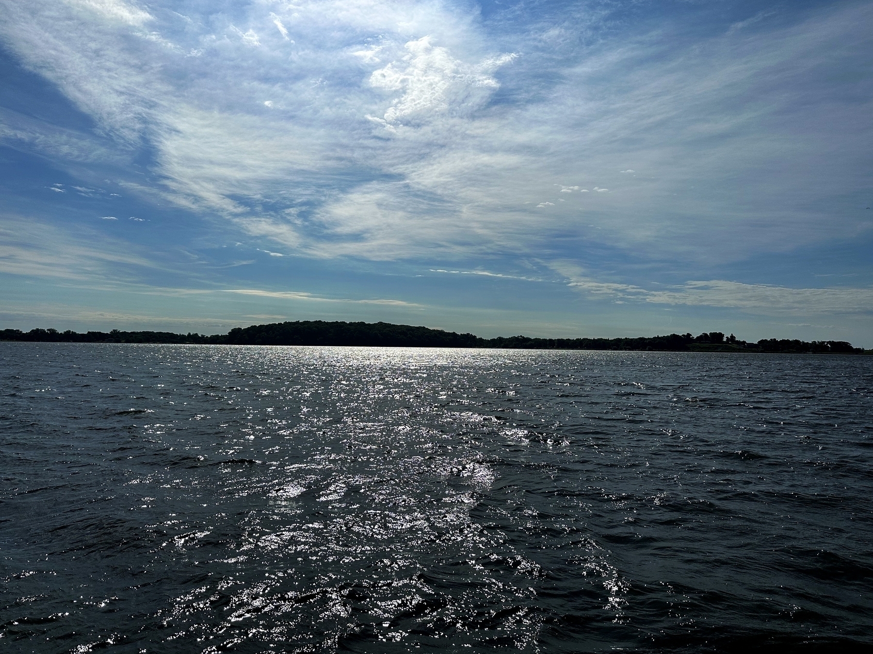 Sunlit waves sparkle on a wide body of water with distant tree-covered land on the horizon under a partly cloudy blue sky.