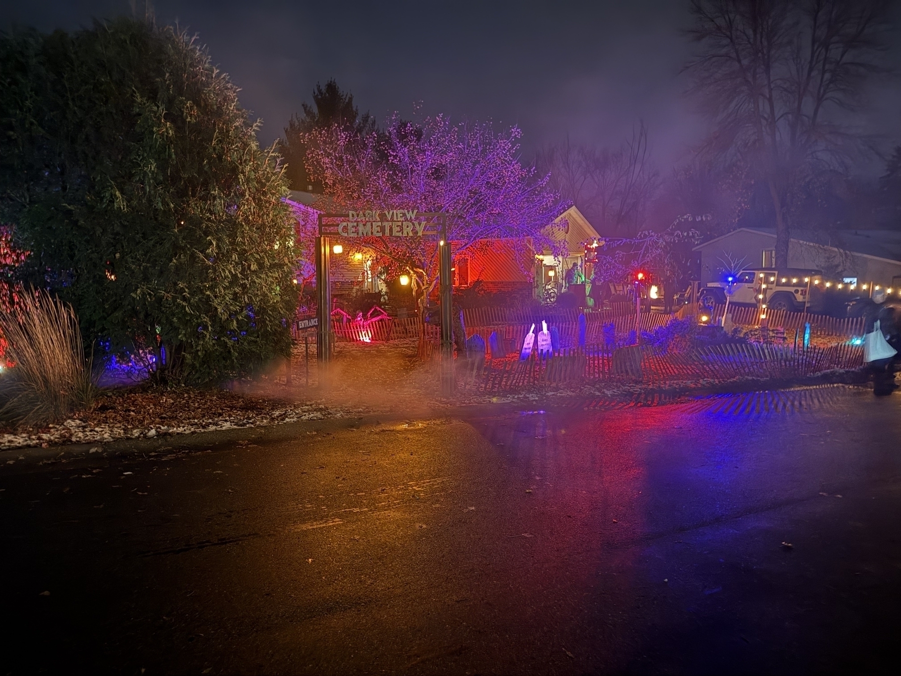 Festive decorations illuminate a yard; gravestones and lights create a spooky, Halloween cemetery scene. A sign reads “Dark View Cemetery Entrance.” Colored lights and fog enhance the eerie atmosphere.