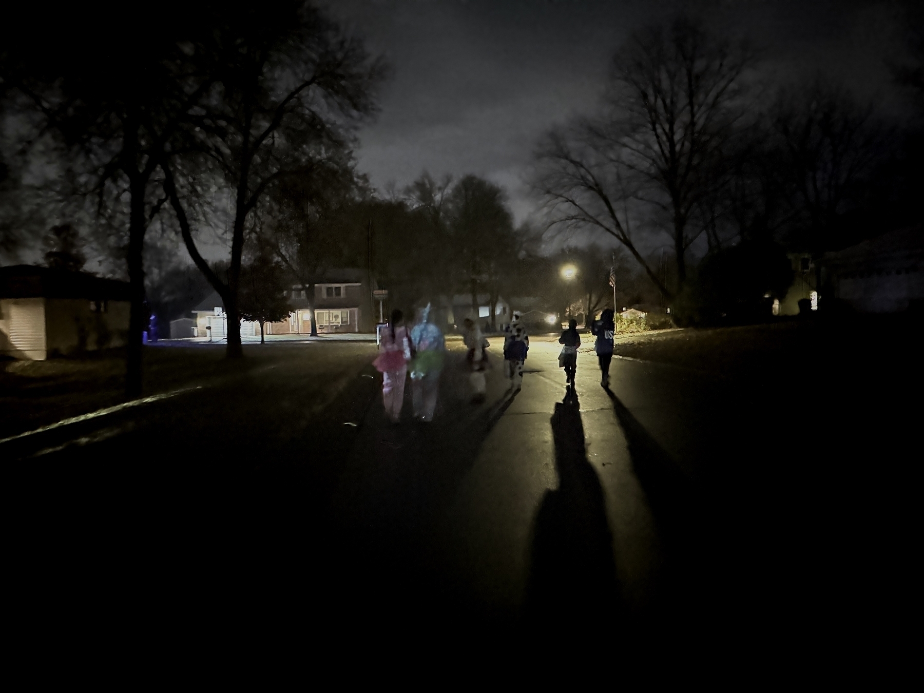 Children walk along a dimly lit street, casting long shadows under streetlights. Leafless trees and residential houses line the quiet suburban neighborhood at night.