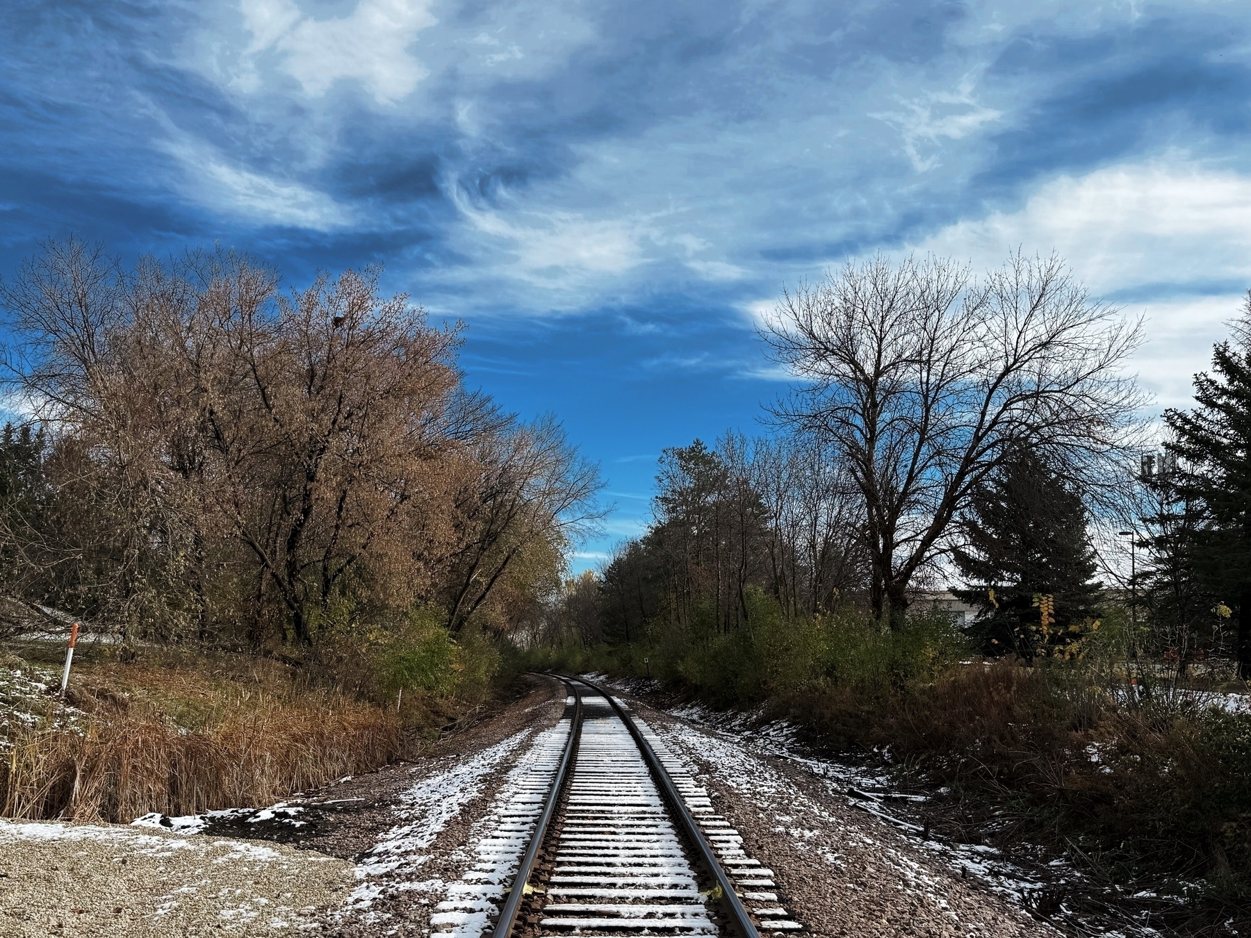 Railroad tracks stretch into the distance, lined with sparse, snow-dusted trees and undergrowth. Overhead, a partly cloudy sky fills the backdrop, creating a serene, rural scene.