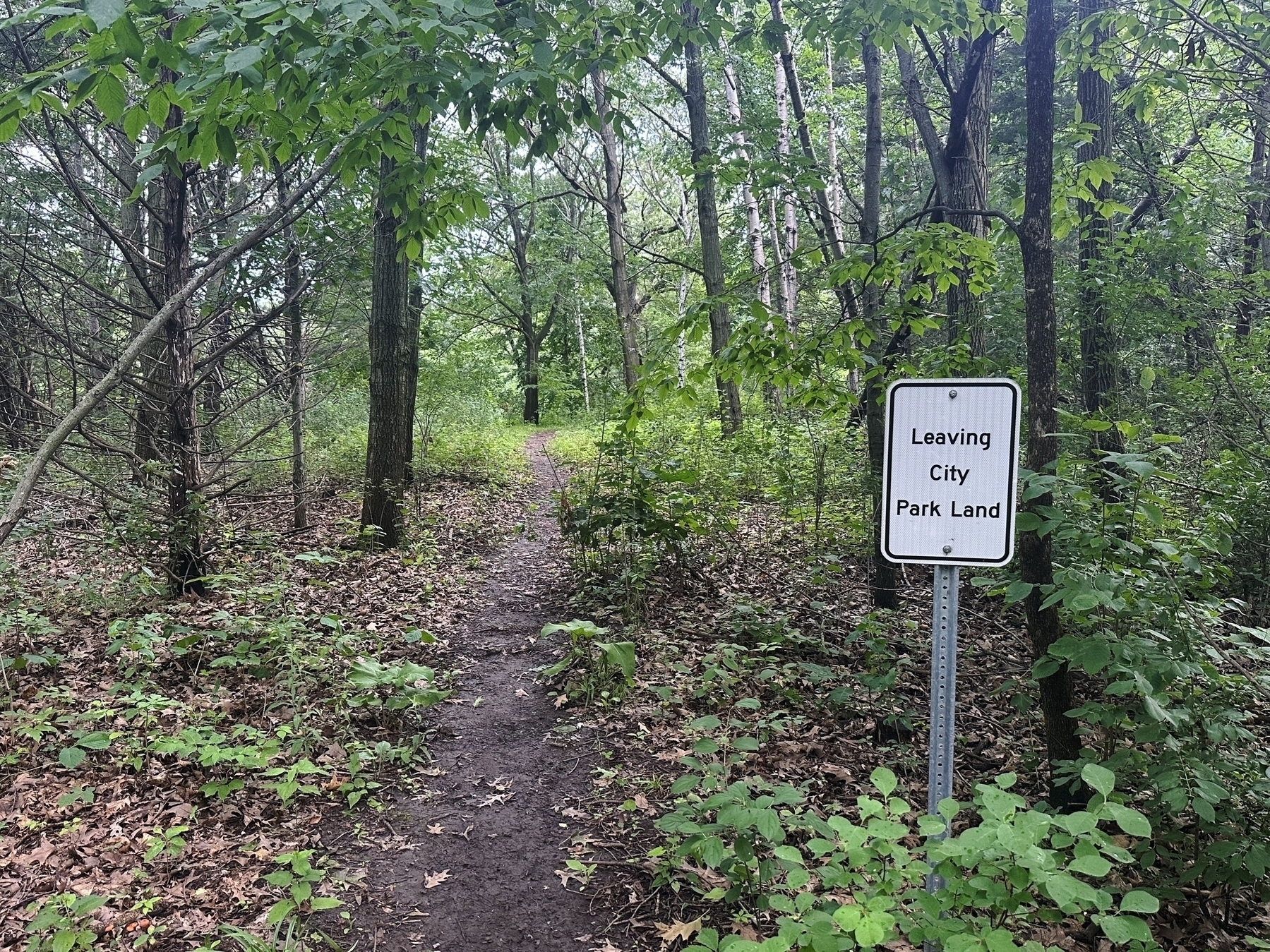 A sign stating Leaving City Park Land stands beside a narrow dirt trail, which winds through a densely wooded area with trees and green undergrowth.