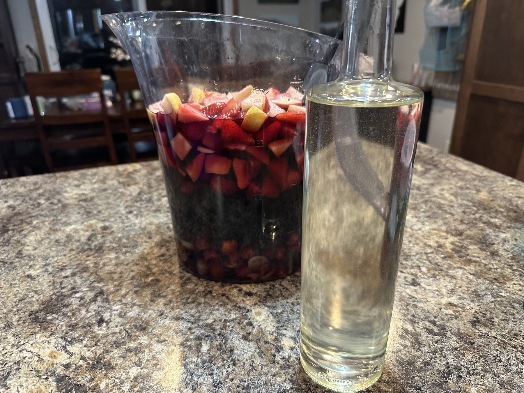 A glass pitcher containing chopped fruits and liquid sits on a marbled countertop next to a clear glass bottle filled with a pale yellow liquid in a kitchen setting.