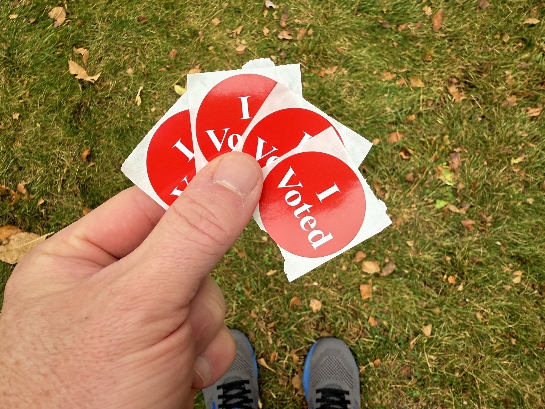 A hand is holding several red I Voted stickers above a grassy background.