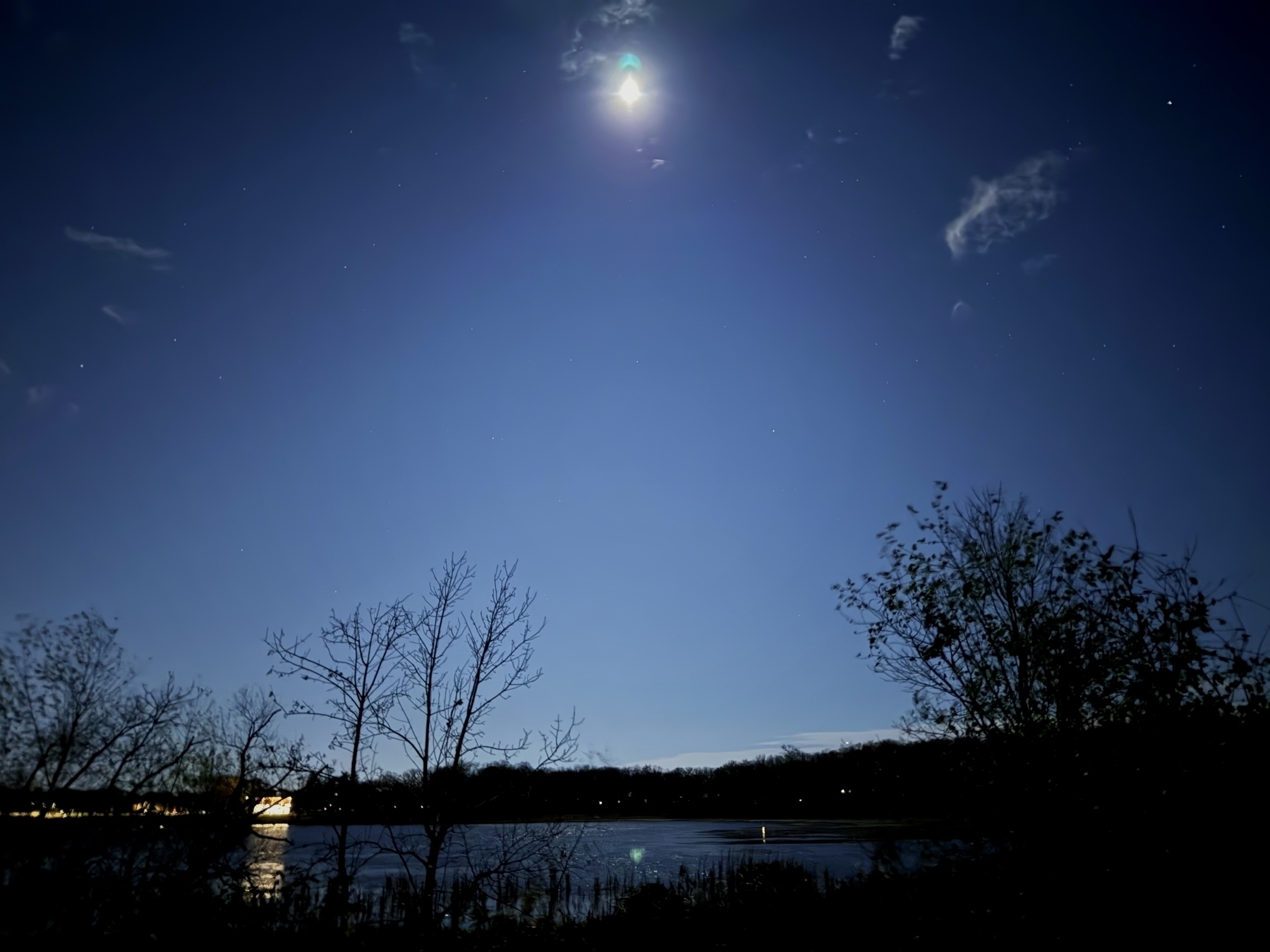 Moon illuminates a dark night sky, casting light over a calm lake. Silhouetted trees and distant shoreline create a serene, natural atmosphere. Clouds and stars dot the expansive sky.