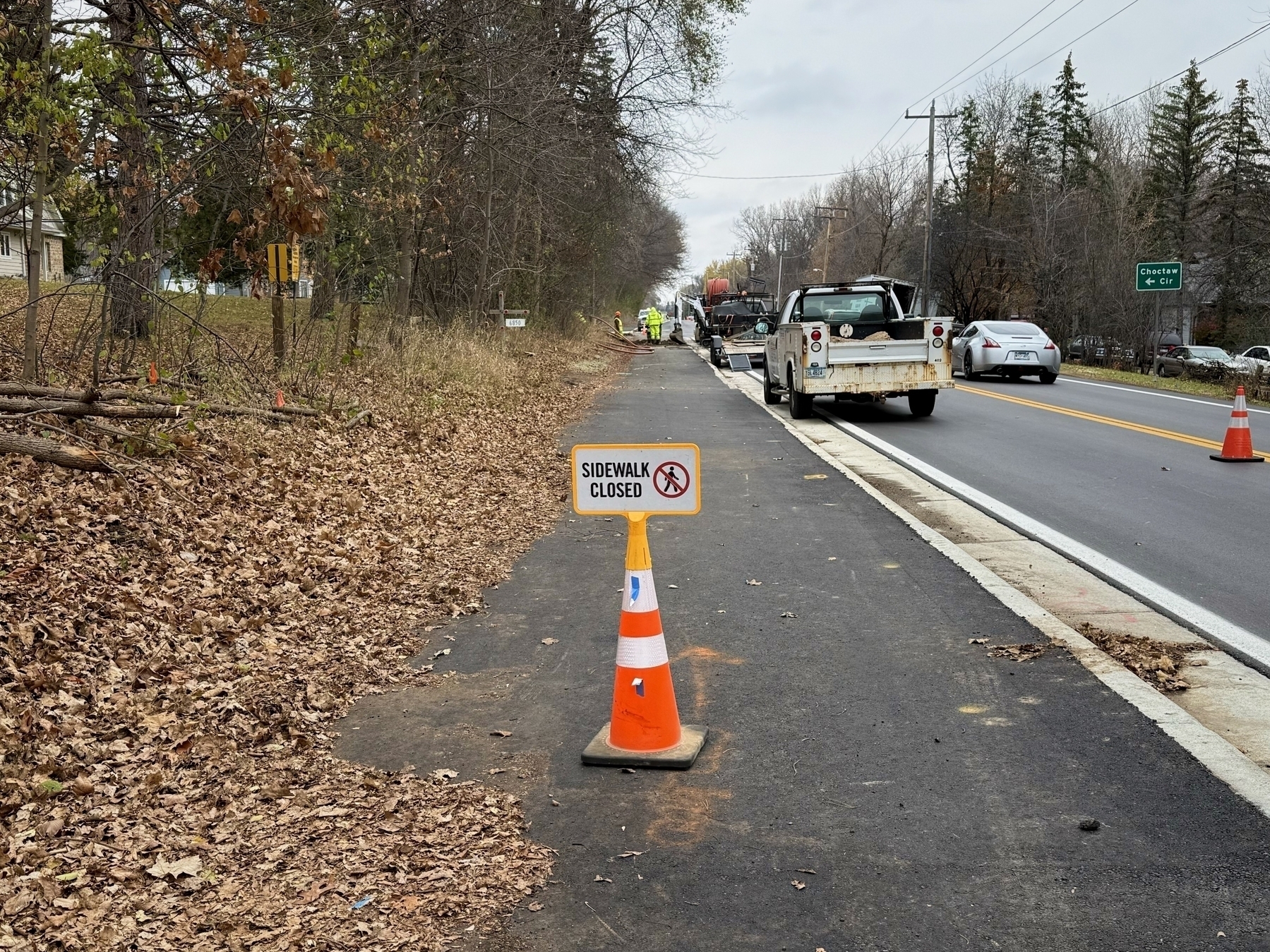 An orange traffic cone with a “SIDEWALK CLOSED” sign blocks a path beside a road, surrounded by fallen leaves and trees, while vehicles and workers continue in the background.