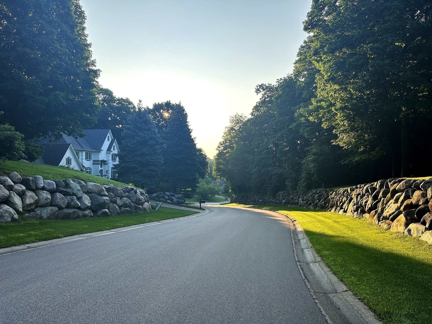 A paved road curves gently between two stone walls with green grass bordered by tall trees and flanked on the left side by a white house nestled among trees.
