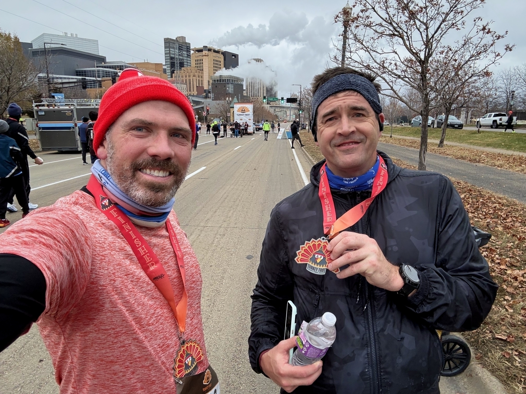 Two men wear medals around their necks after completing a race, standing on a city street with a cloudy sky and bare trees. One holds a water bottle and phone. “FINISHER” is written on the medals.