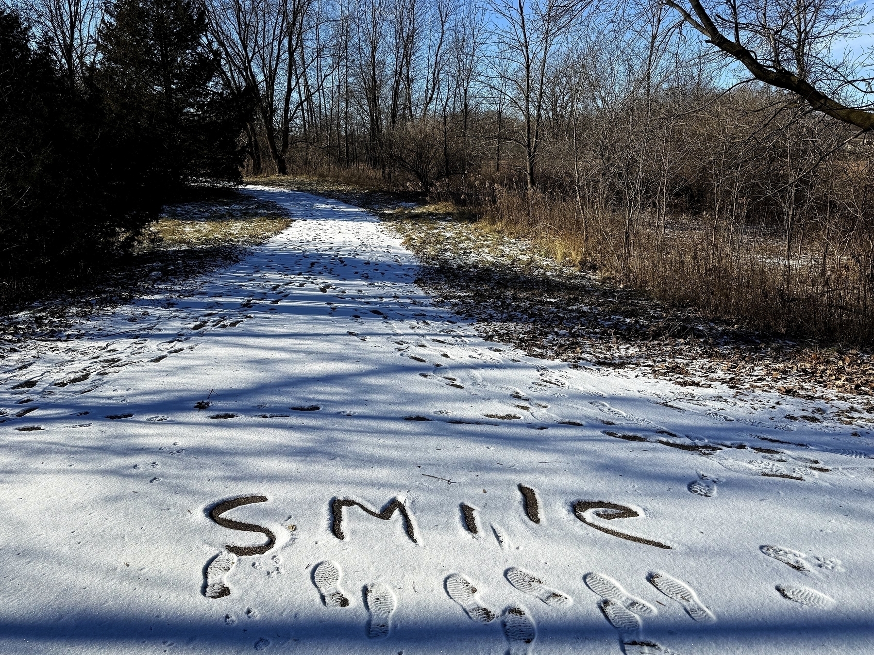 Footprints are scattered over a light dusting of snow on a wooded path, with the word “SMILE” etched into the snow amidst bare trees.