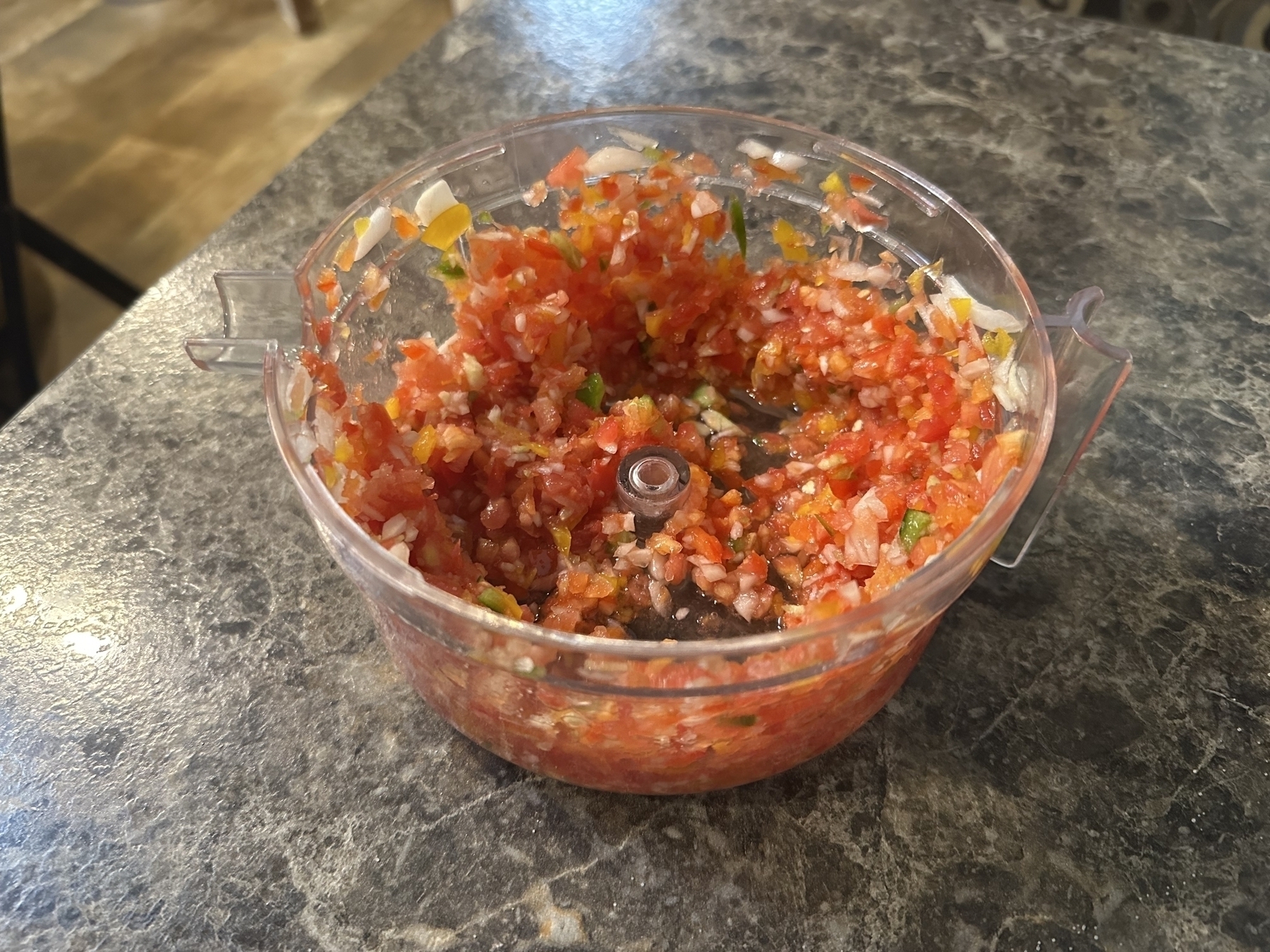 Chopped vegetables (tomatoes, onions, peppers) sit inside a transparent food processor bowl on a dark marbled countertop in a kitchen setting.