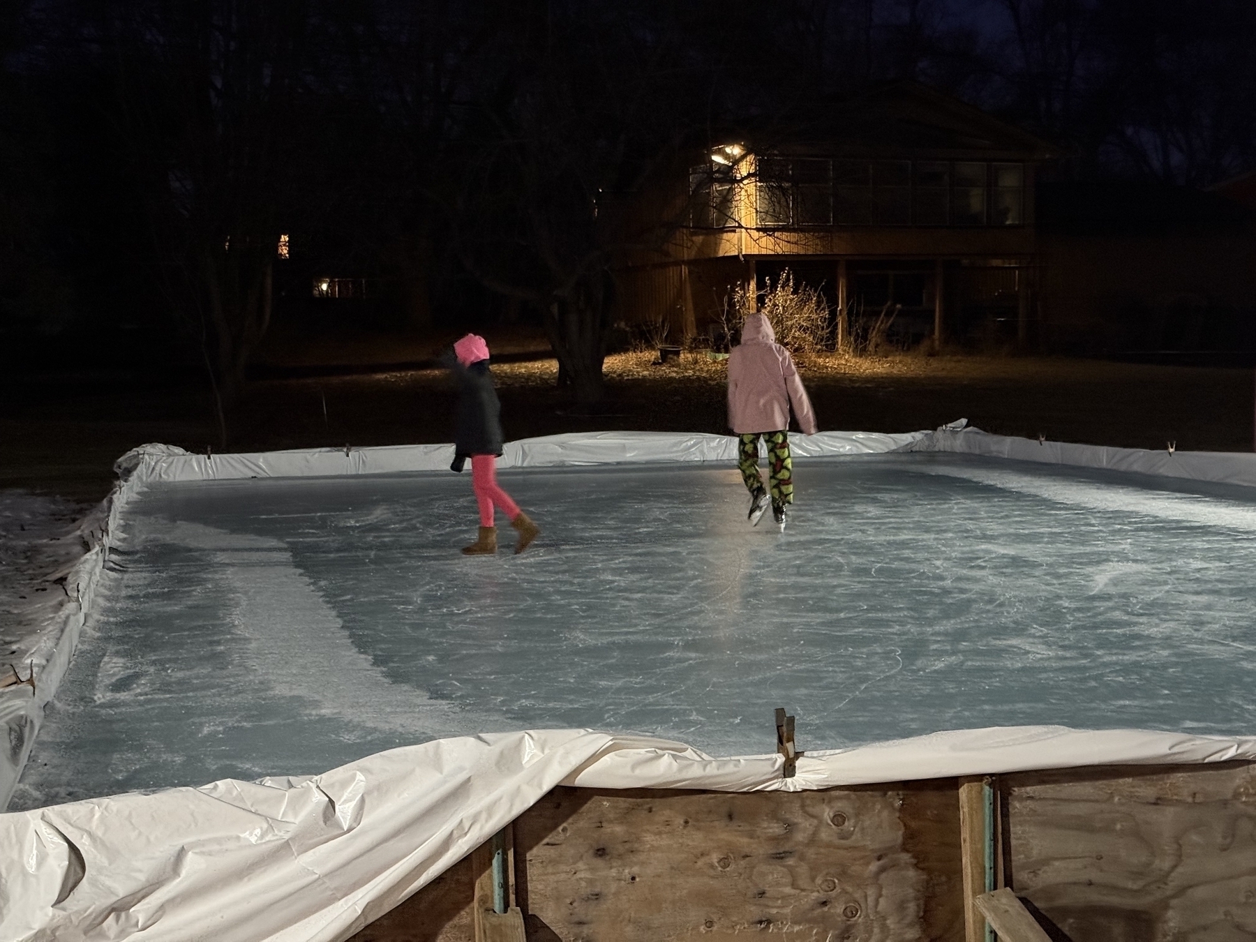 Two people skate on an illuminated outdoor ice rink, situated in a dark backyard environment with house lights in the background. They wear warm clothing, including jackets and hats.