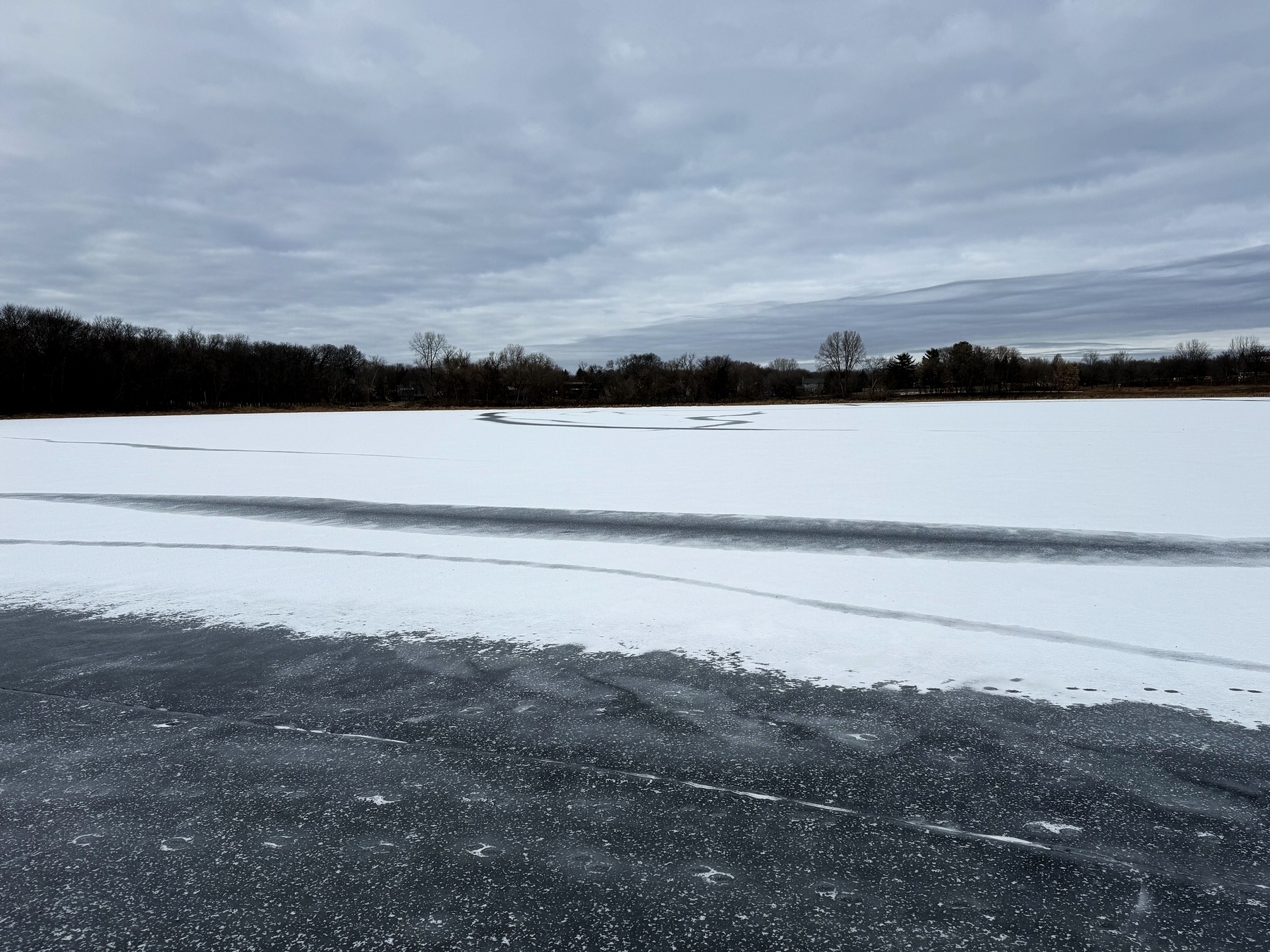 Frozen lake with patches of snow and ice, surrounded by bare trees under a cloudy sky, creating a wintry, tranquil scene.