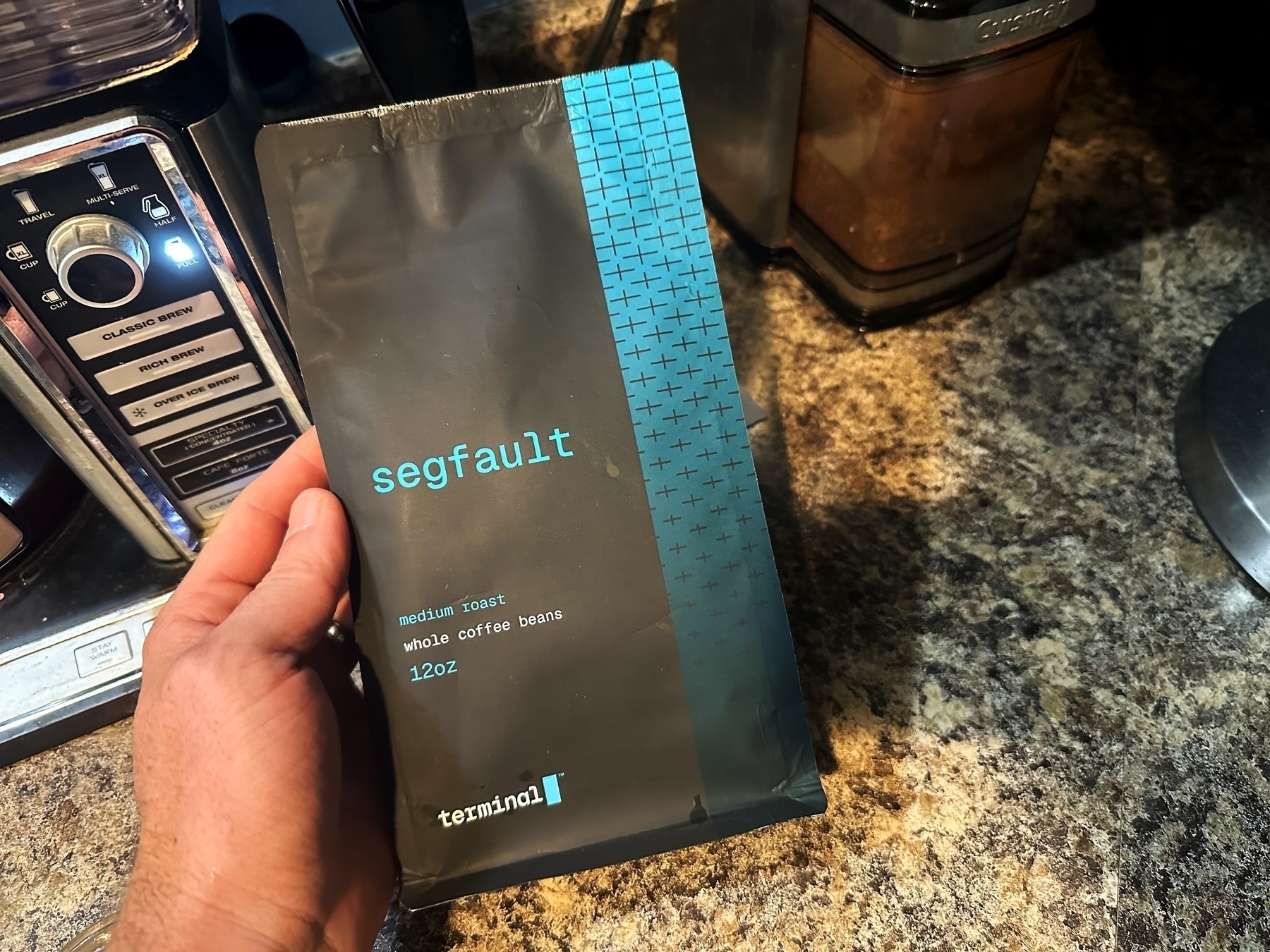 A hand holds a segfault medium roast coffee bag beside a coffee maker on a kitchen counter. The bag is black with light blue text and patterns.