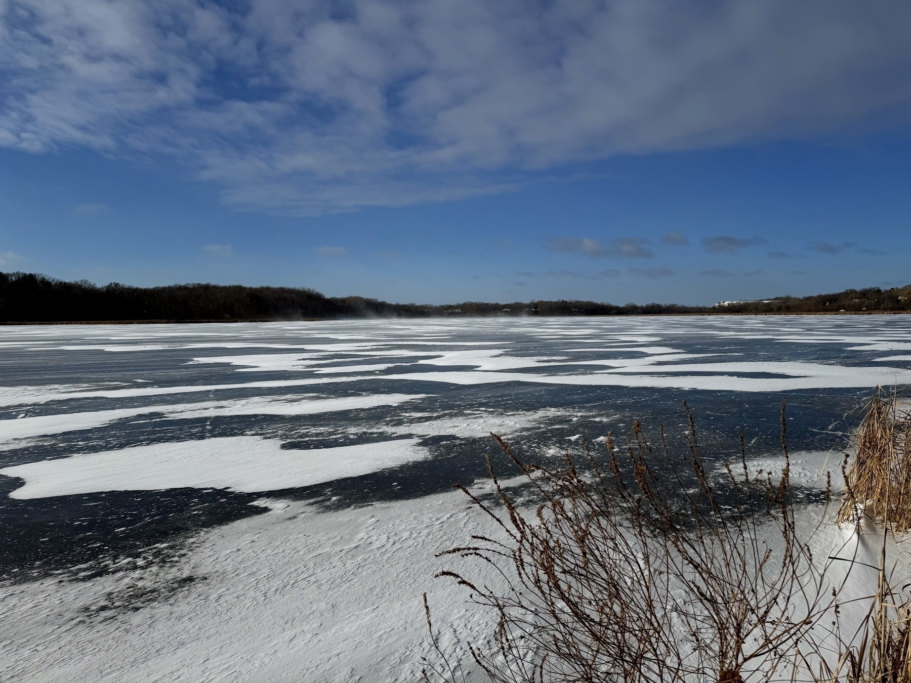 Snow-dusted frozen lake contrasts with patches of dark ice, while dried grass borders the foreground under a partly cloudy blue sky, capturing a serene winter scene.