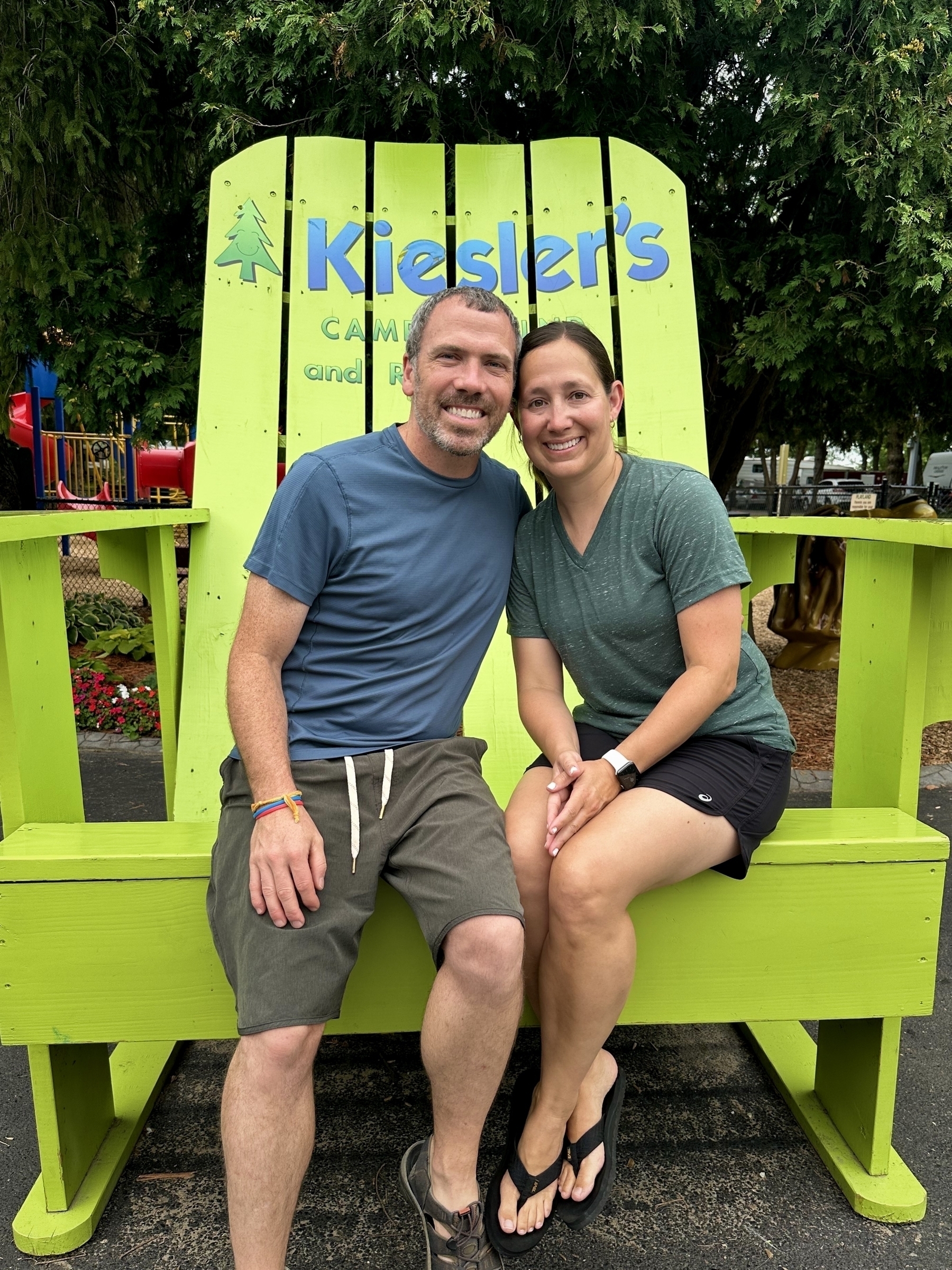 Two people are sitting and smiling on a large, bright green wooden chair. Surrounding them are trees and colorful playground equipment. The text on the chair reads, “Kiesler’s Campground and RV Resort.