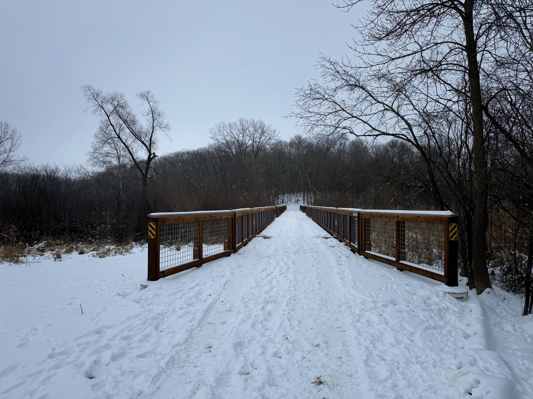 A snow-covered wooden bridge extends into a forest of bare trees under an overcast sky, creating a serene winter scene. Bright yellow warning stripes are visible on the bridge’s railings.