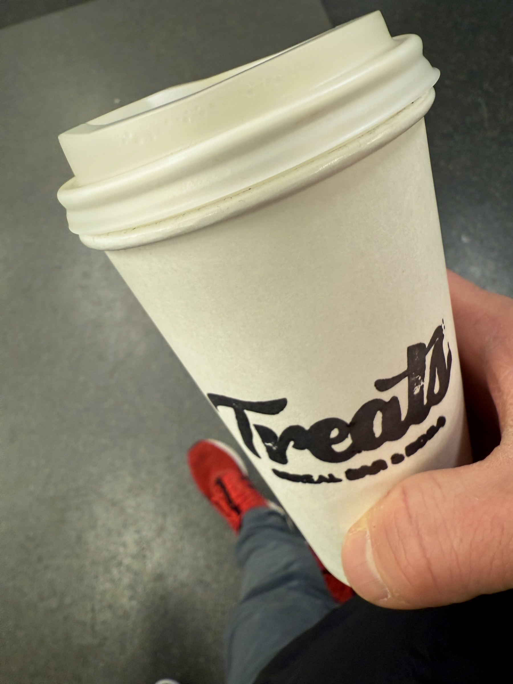 A hand holds a white coffee cup labeled “Treats” in bold, black font, against a blurred indoor background. Red shoes and gray pants are partially visible below.