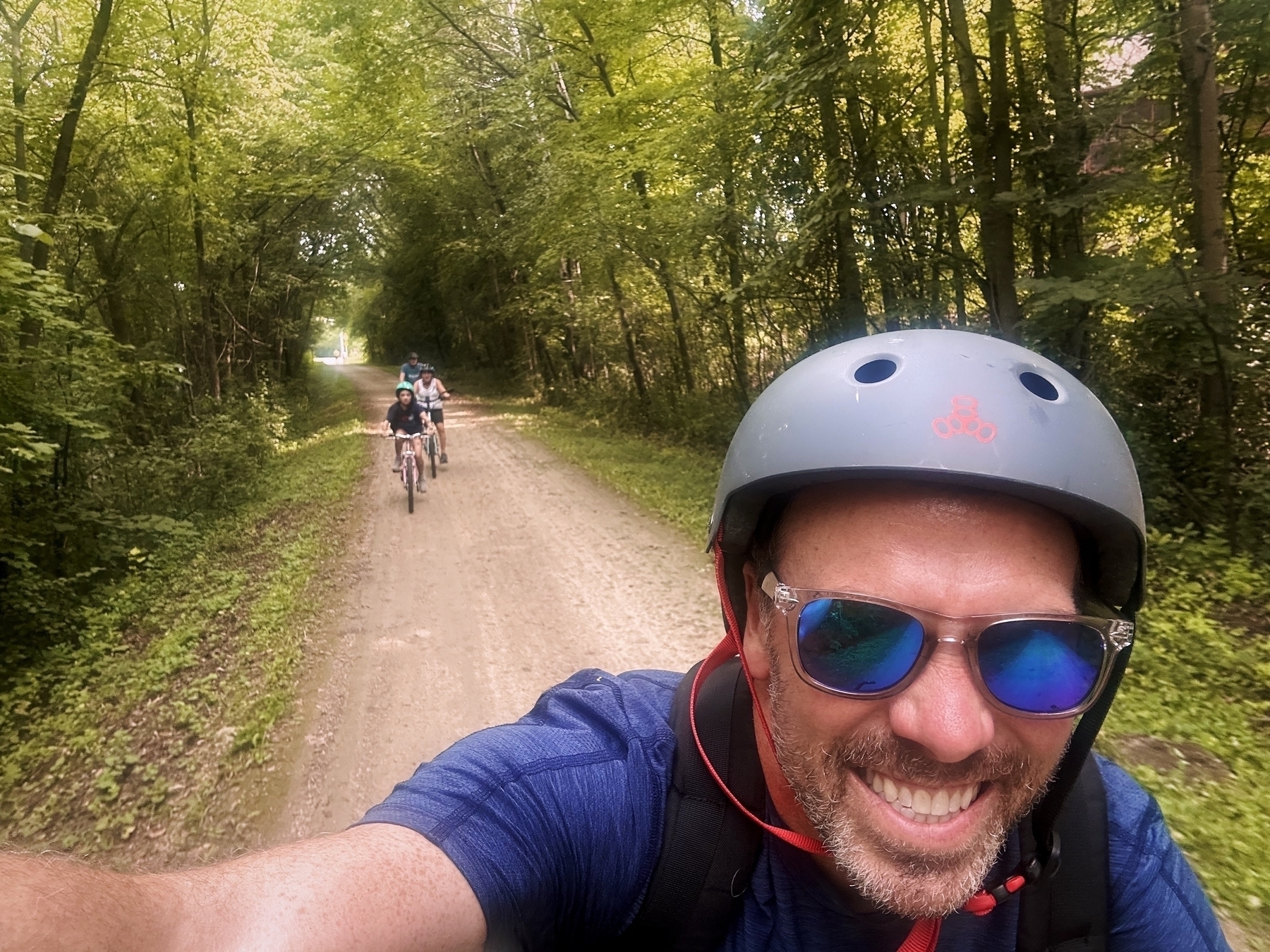 A man wearing a helmet and sunglasses is riding a bicycle and smiling on a forested dirt trail, followed by two more cyclists. Green trees line the path closely on both sides.